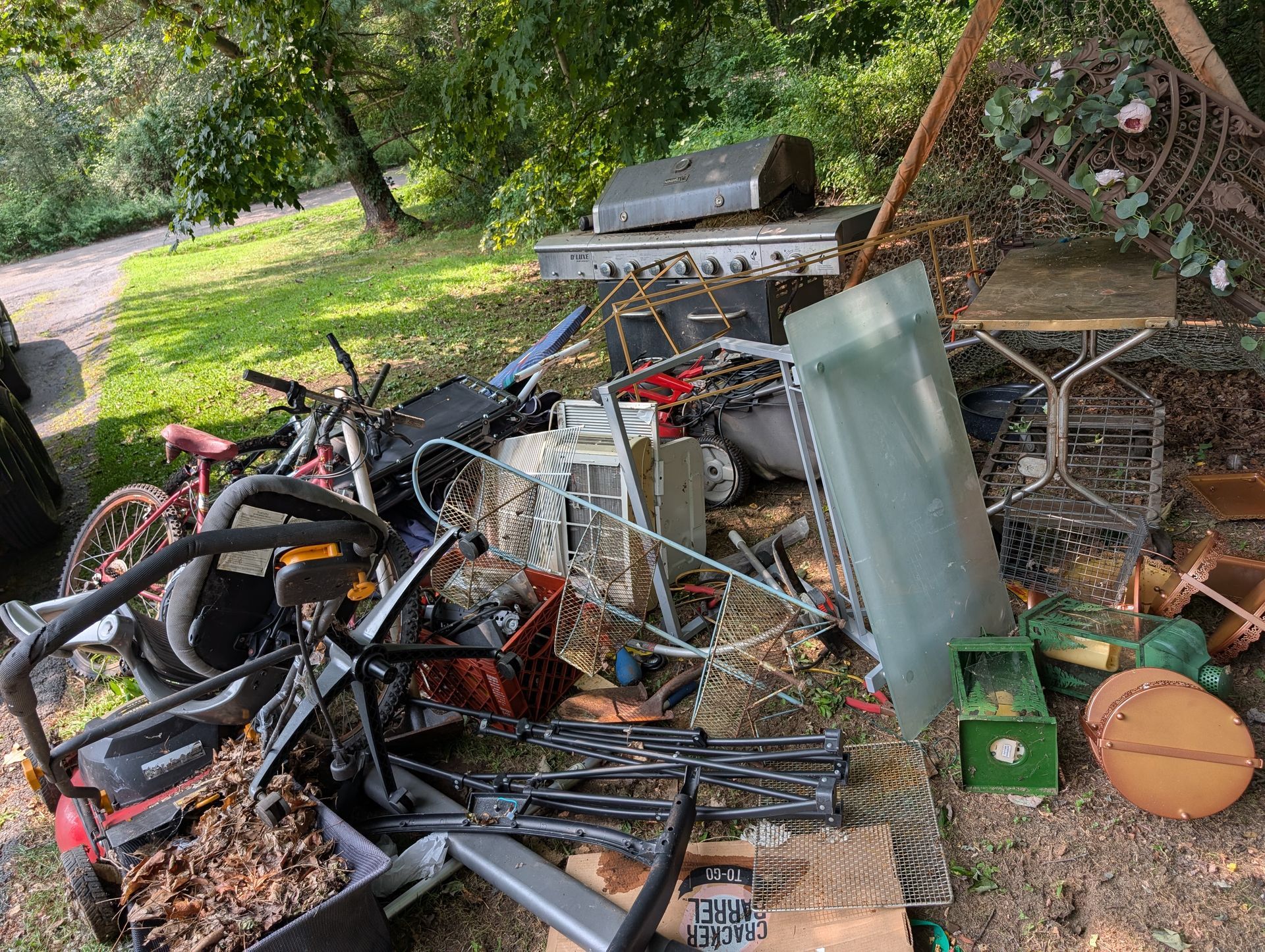 Pile of discarded items, including a grill, bike, lawnmower, and various metal and plastic objects, on a grassy area.
