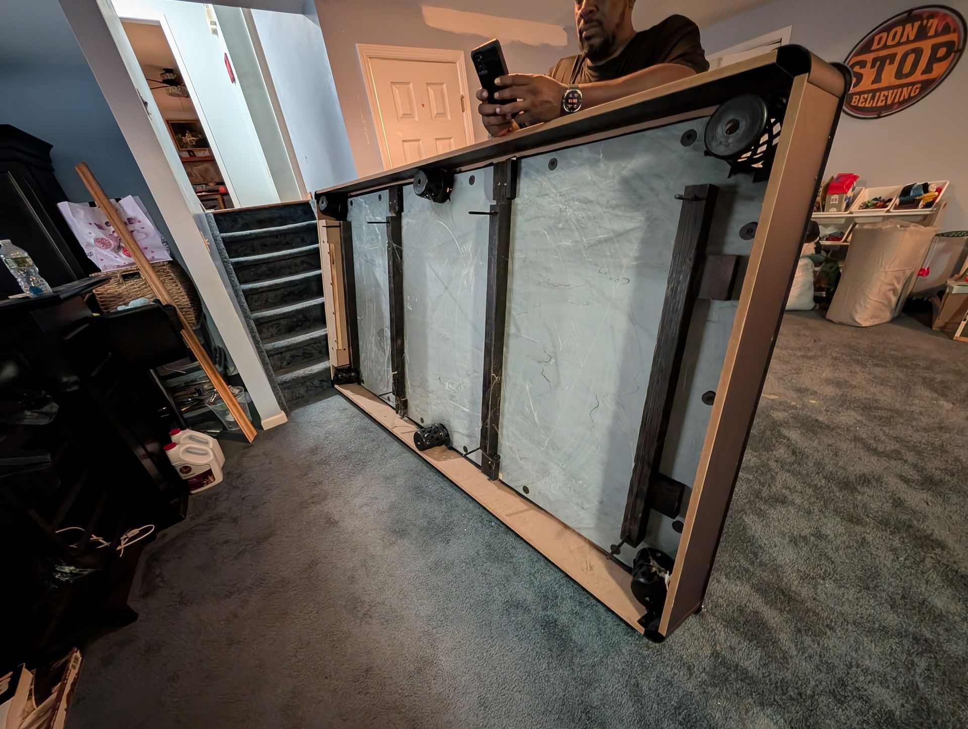 Person holding a phone, inspecting the underside of a light-colored pool table. The table is on a gray carpet.