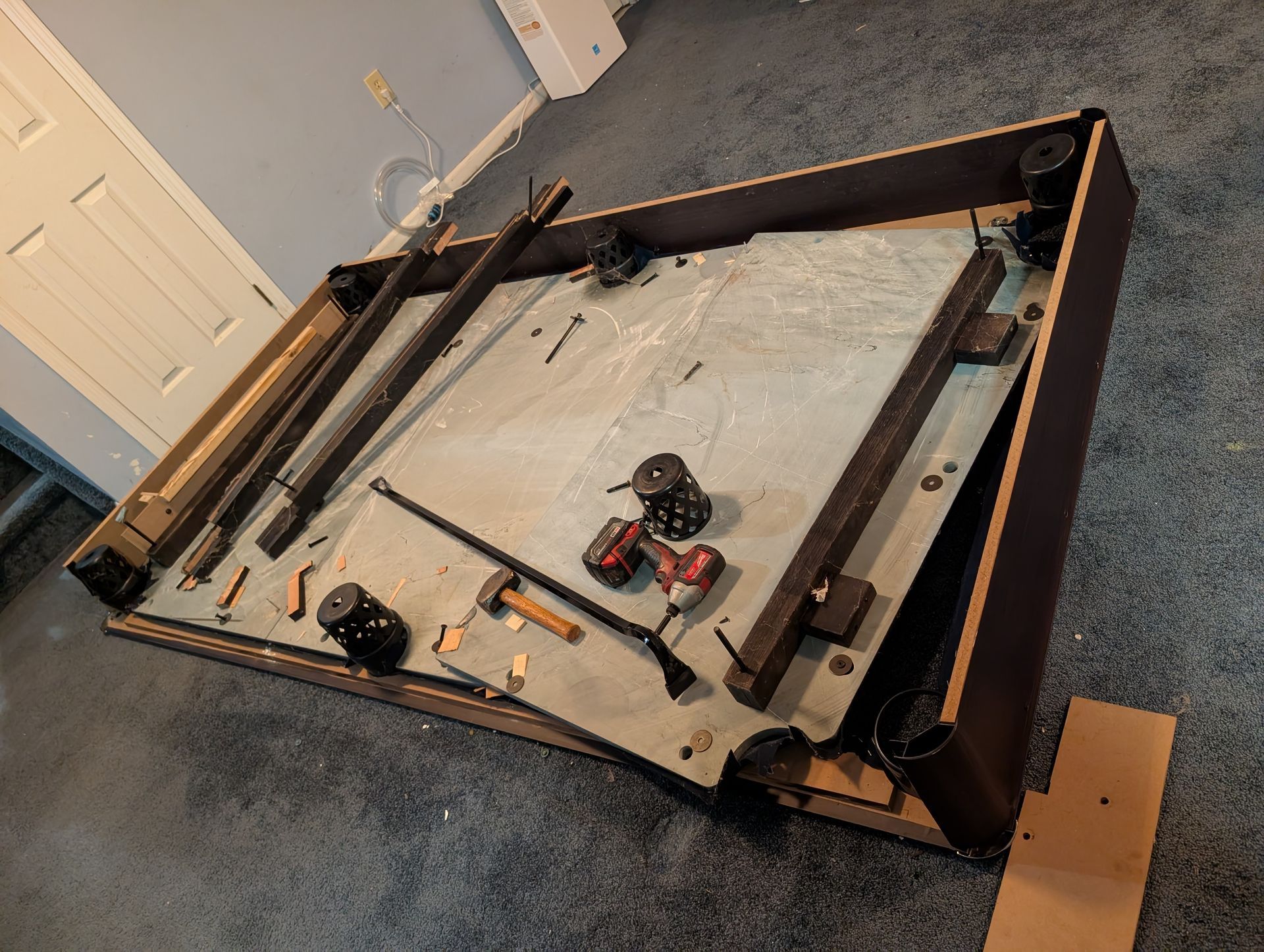 Disassembled pool table on the floor, parts and tools scattered. Brown wood frame, gray playing surface, tan carpet.