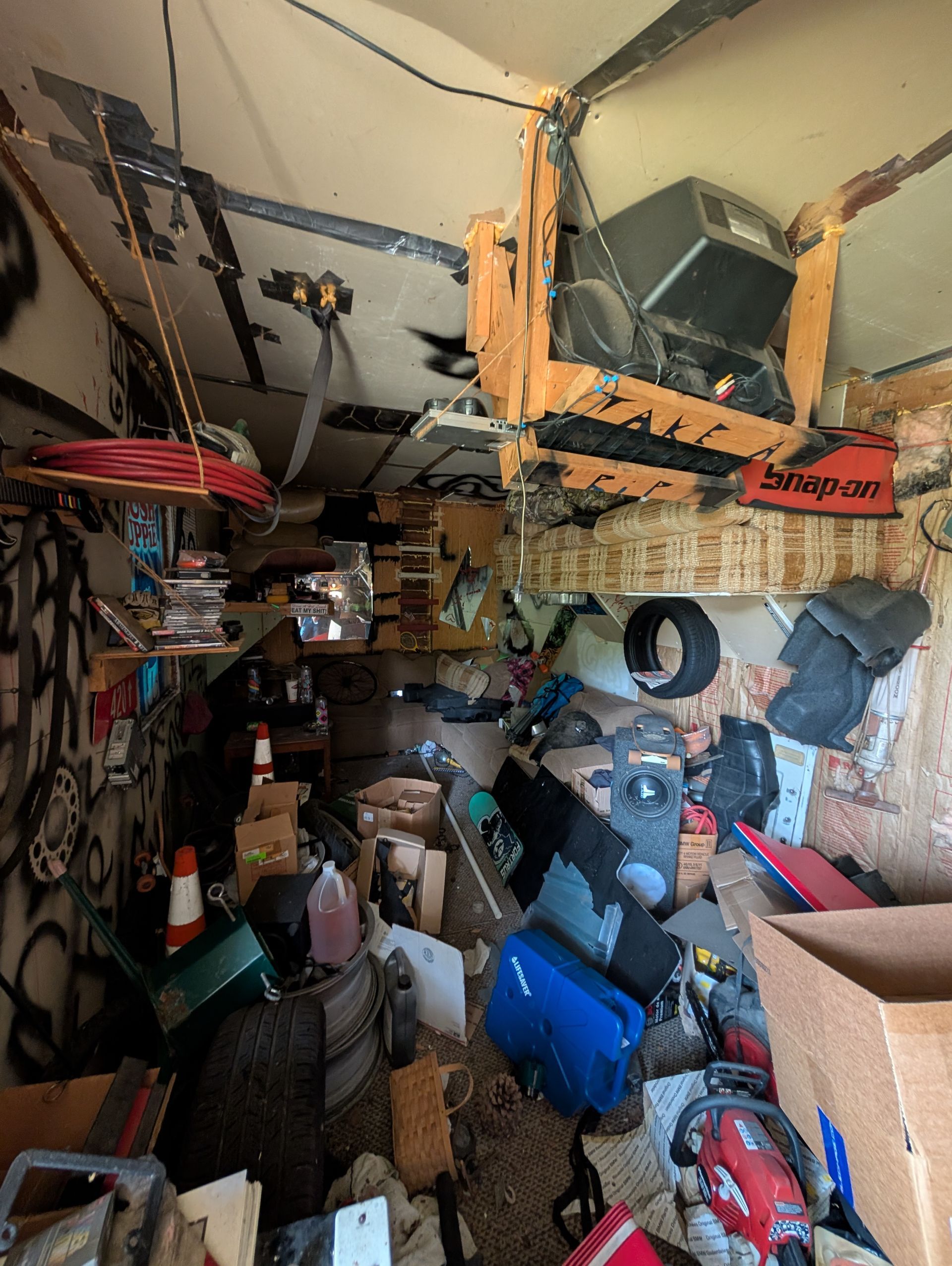 Messy, cluttered interior of a structure. Items piled everywhere, including tires, boxes, and electronic equipment. Graffiti on walls.