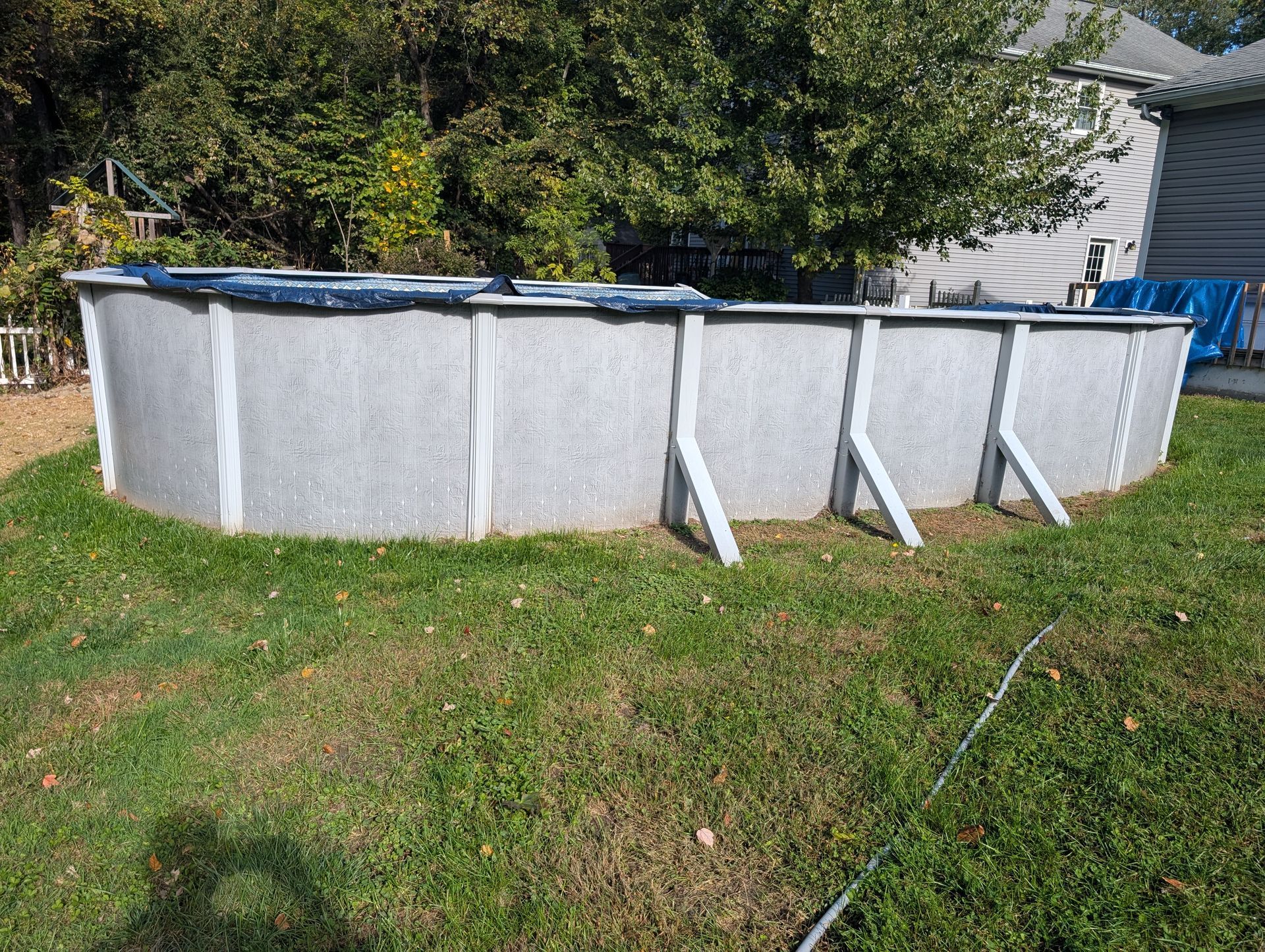 An above-ground pool with a dark cover sits in a grassy yard, trees and a house in the background.