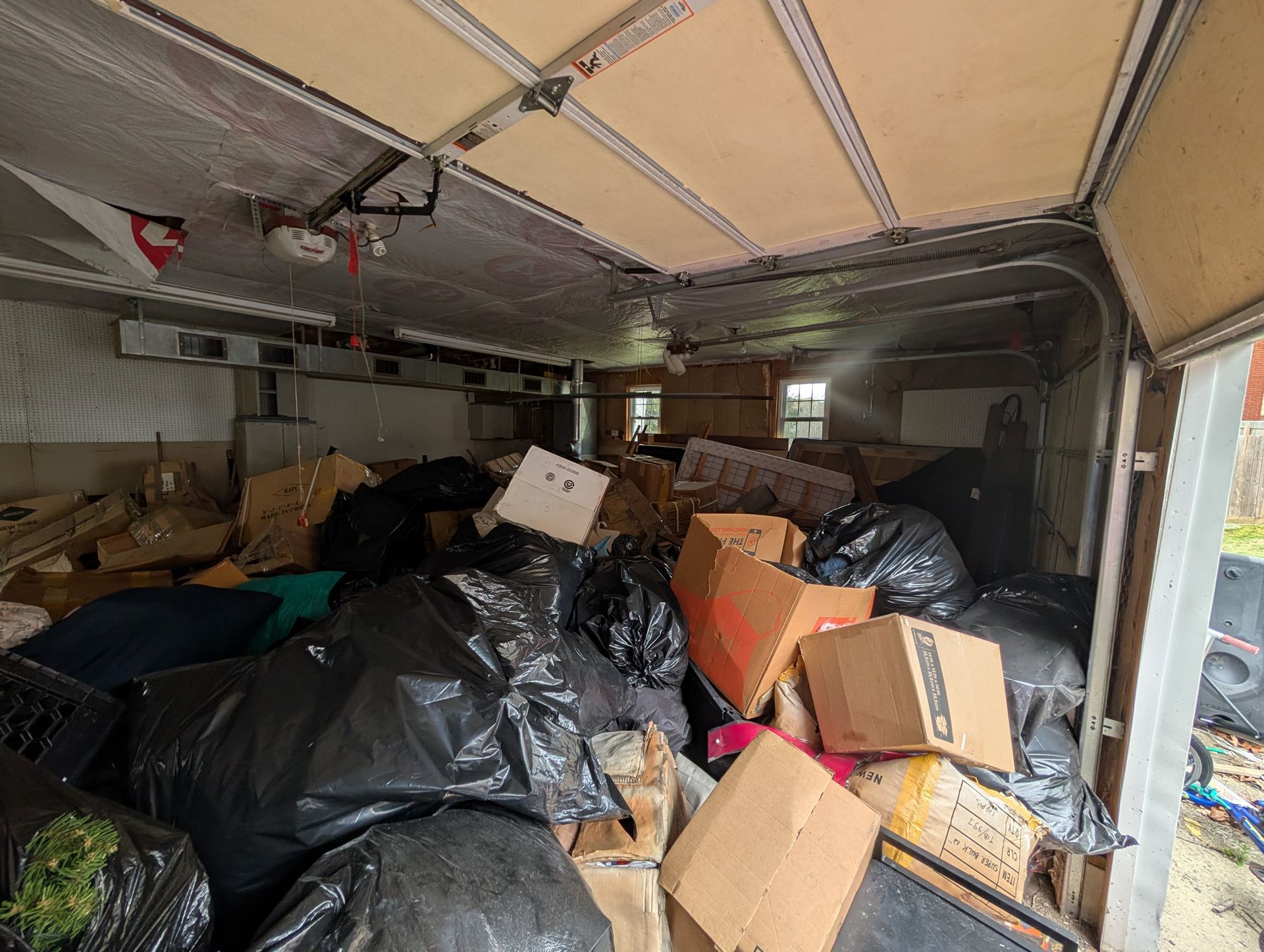 Cluttered garage filled with black trash bags and cardboard boxes. Open garage door.