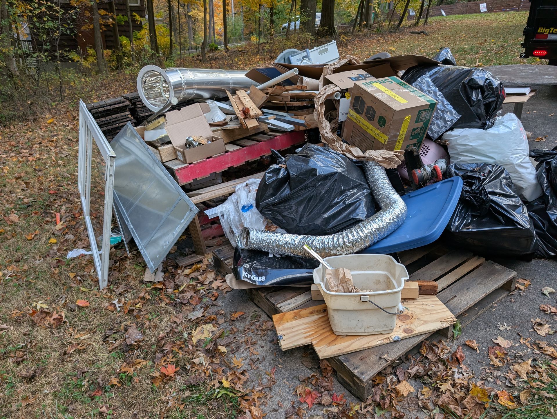 Pile of trash and debris including pallets, boxes, and a bucket, outdoors on fallen leaves.