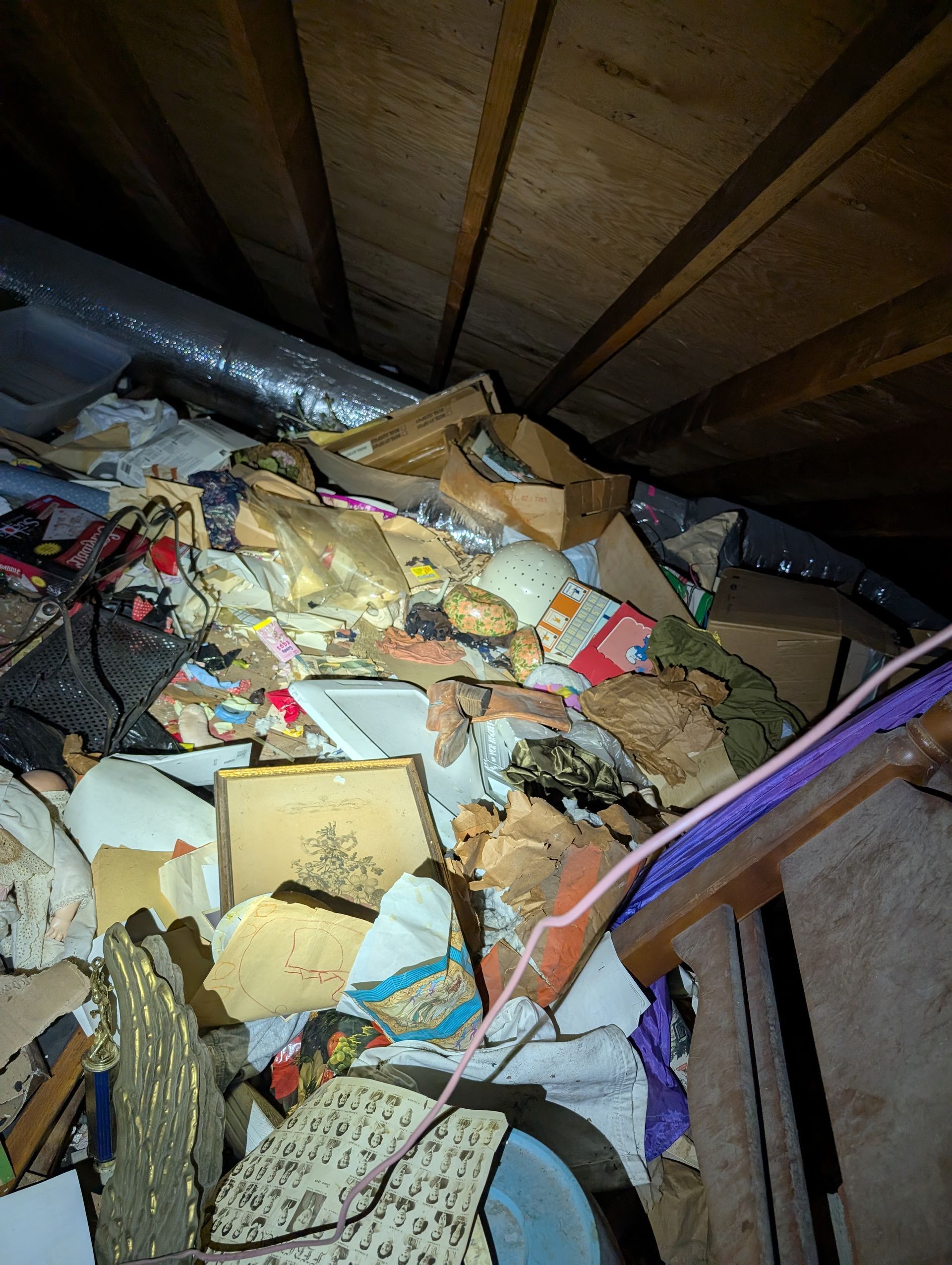 Pile of cluttered items in an attic, including boxes, trash, and insulation. Dark, enclosed space.