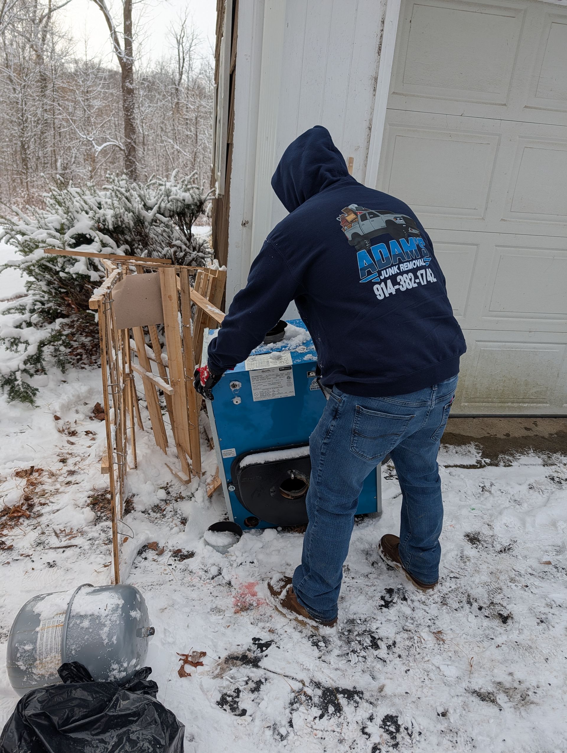 Person in a hooded sweatshirt near a generator in snow. Gray garage door and snow-covered foliage are visible.
