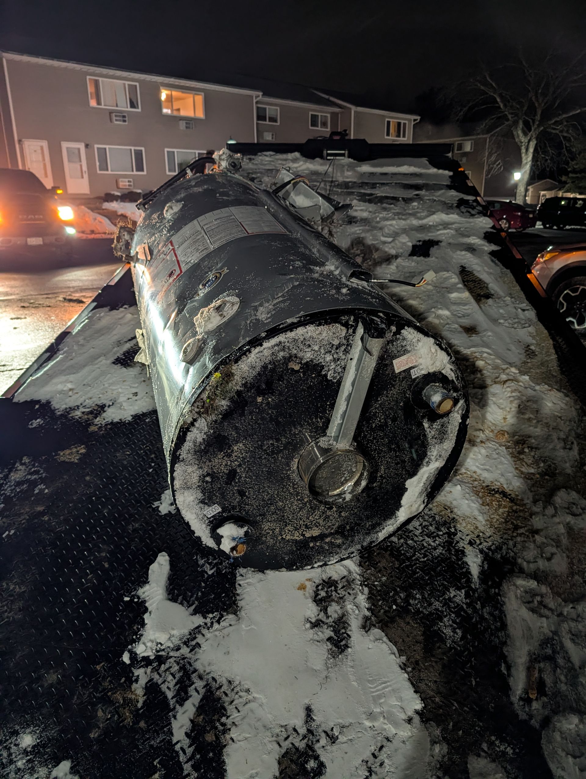 Black water heater on snow-covered surface. Dark, nighttime scene; residential area in the background.