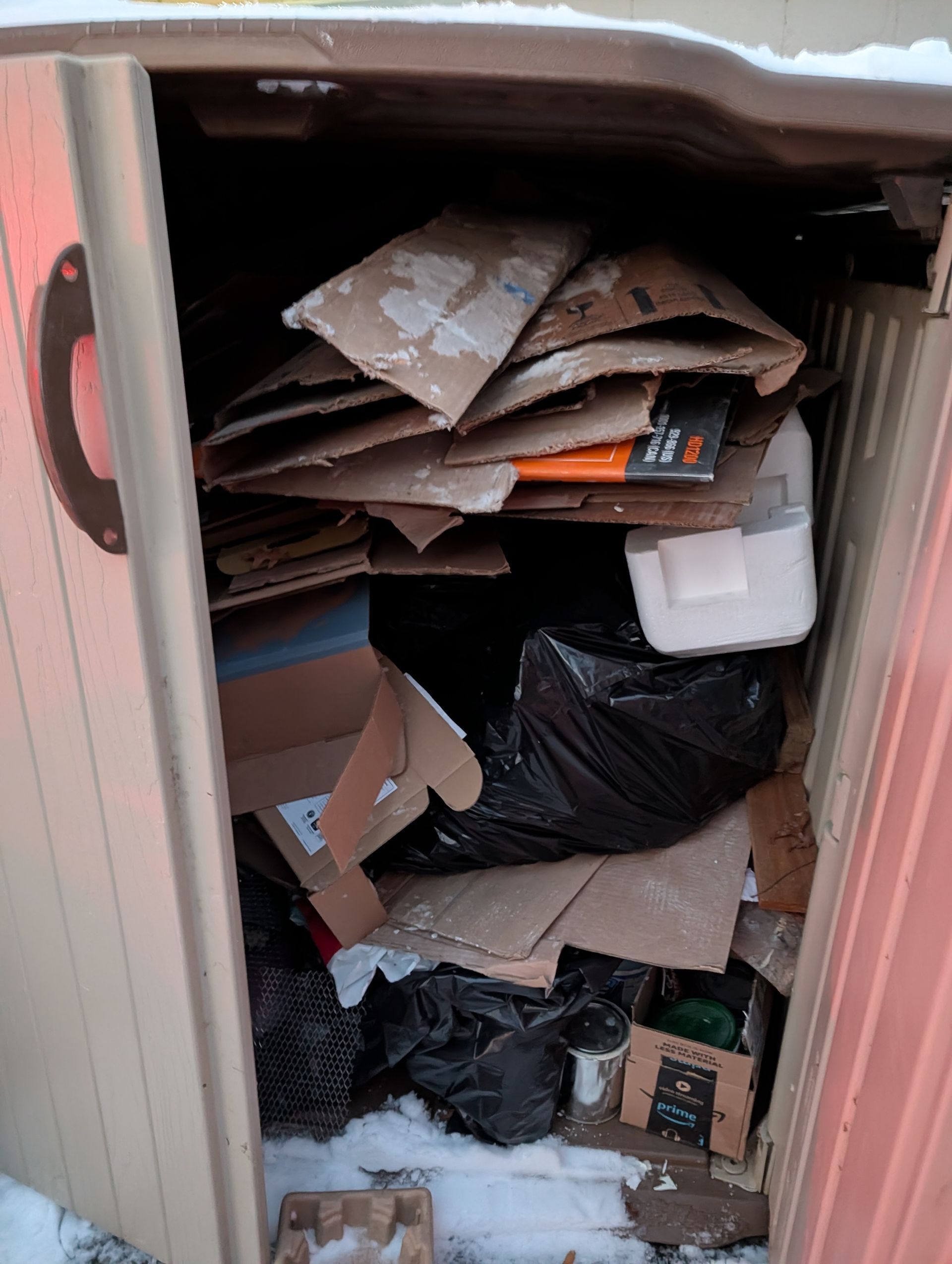 A cluttered outdoor storage shed with cardboard boxes, trash bags, and other items.