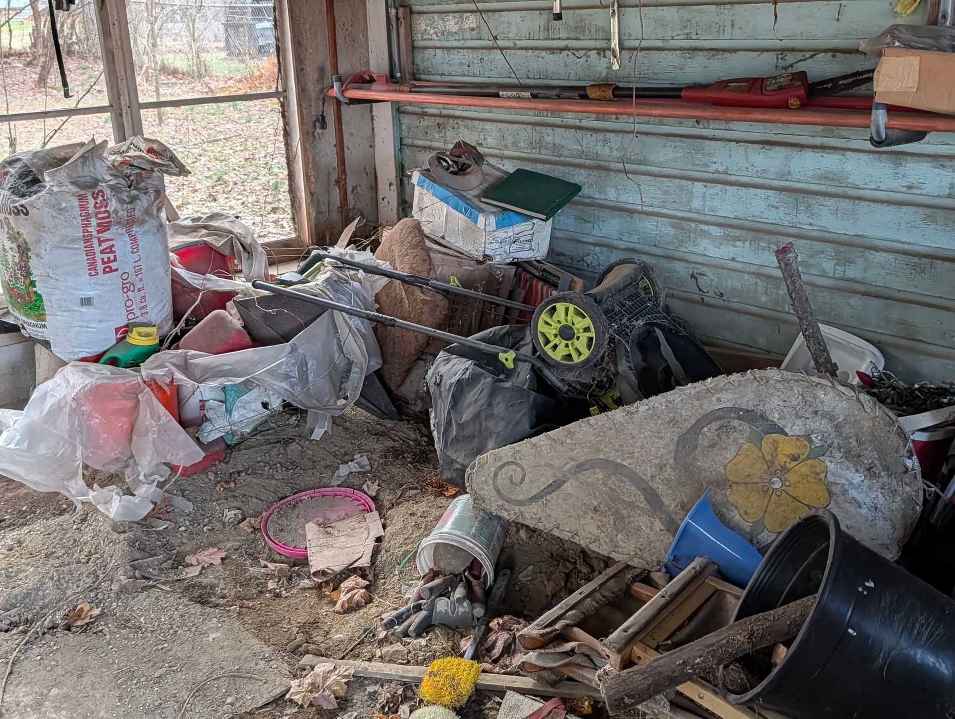 Cluttered shed interior: trash bags, lawn mower, debris on floor, weathered wood walls, various tools and objects.