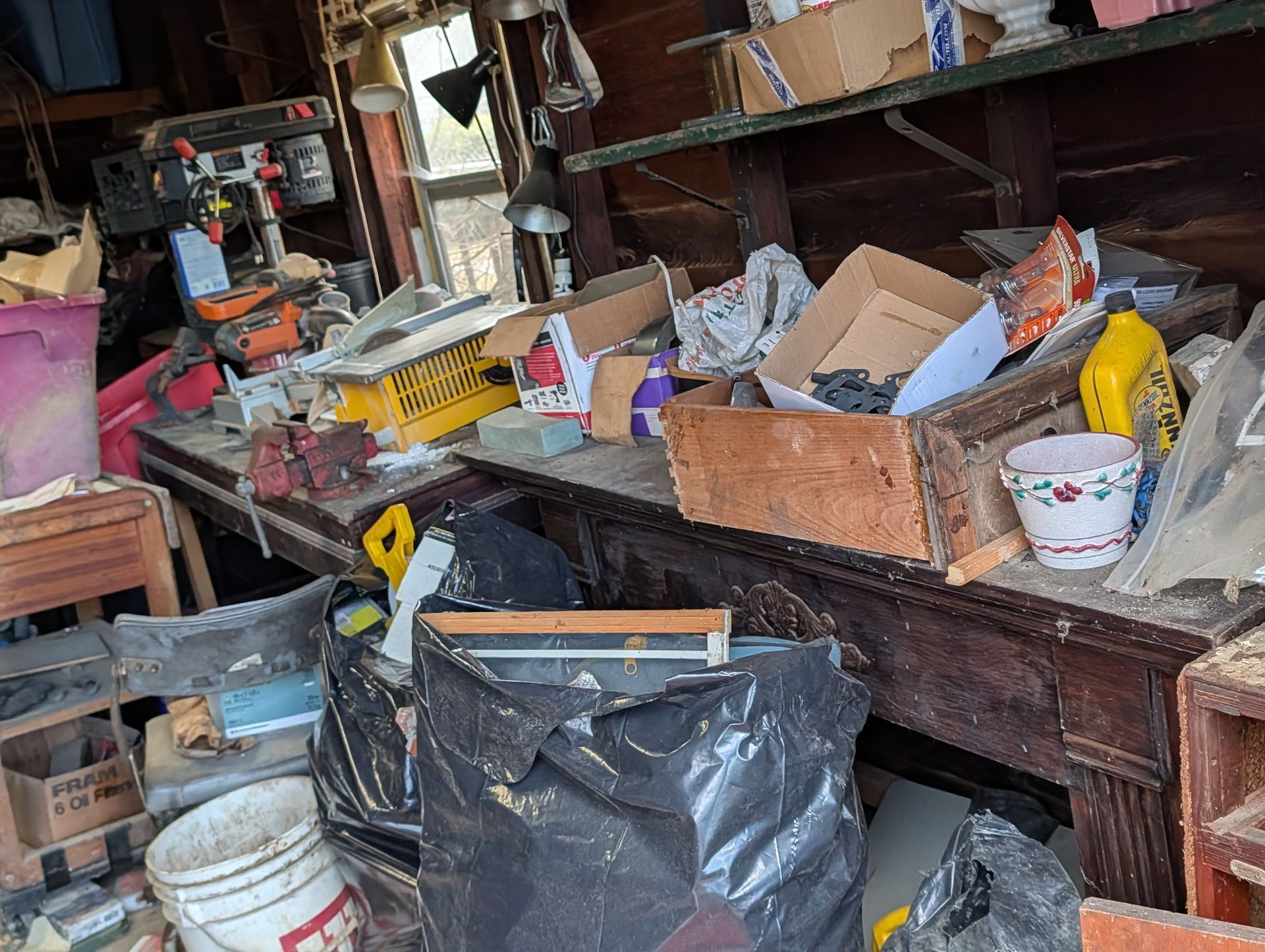 Cluttered workbench in a garage with tools, boxes, and a trash bag.