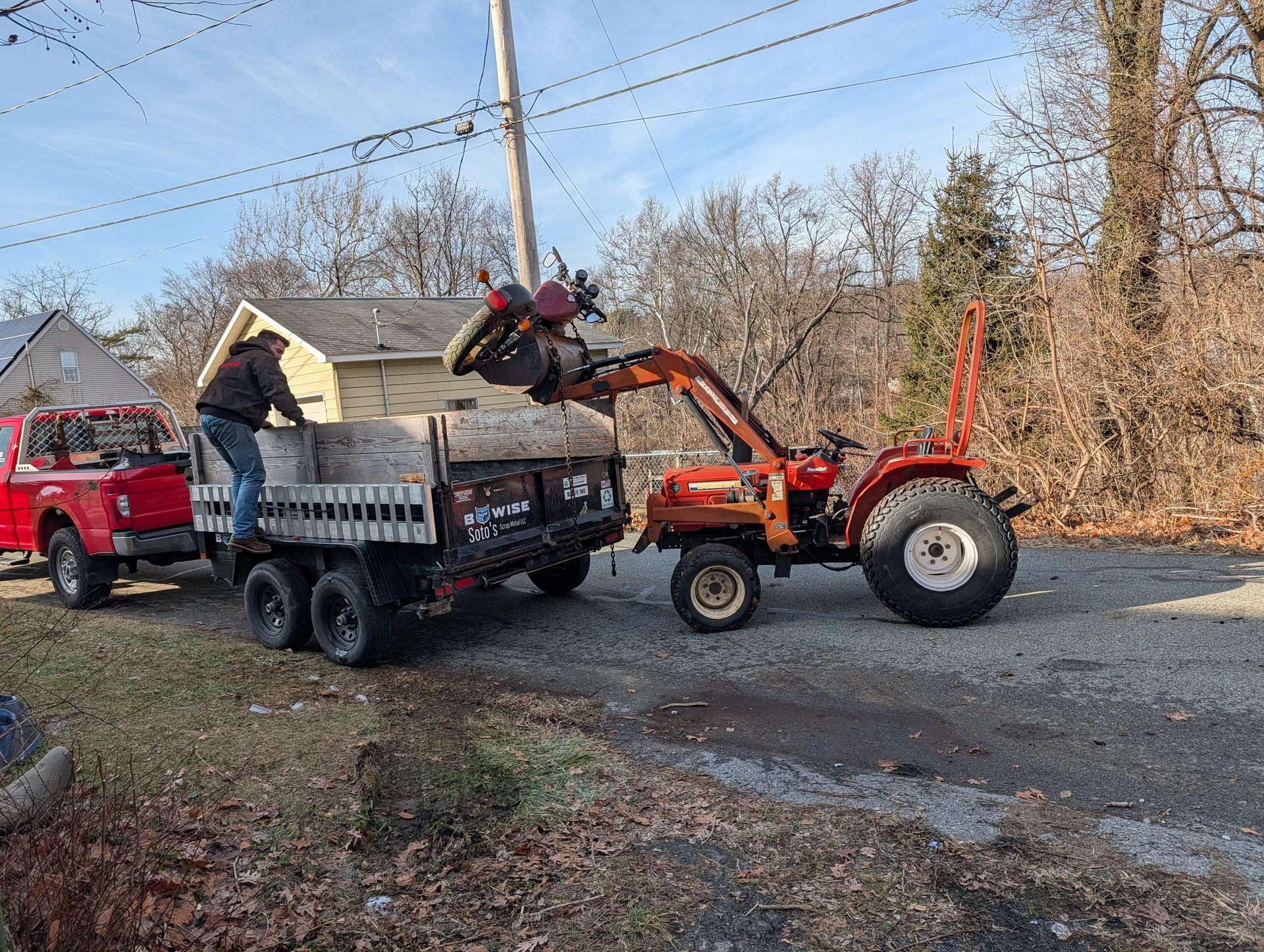 Man loads debris from tractor into a dump trailer parked on a gravel drive. A red Jeep is beside the trailer.