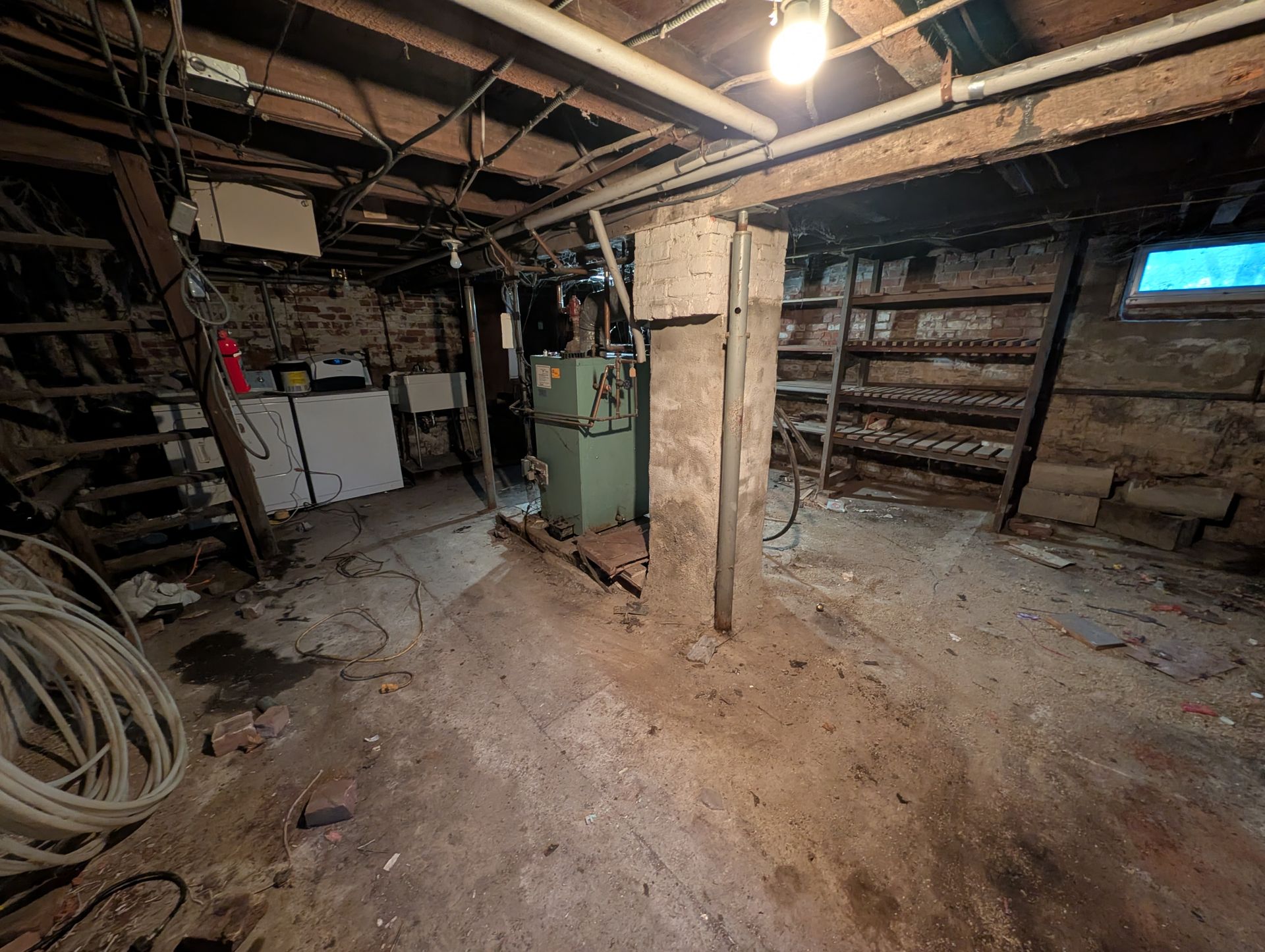 Cluttered basement with exposed beams, brick walls, and concrete columns. Contains a furnace, shelves, and a washing machine.