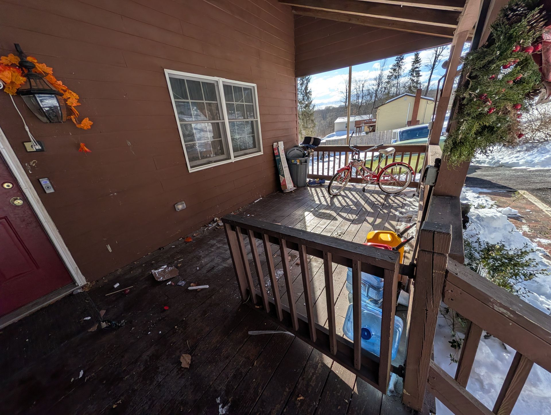 Brown porch with railing, door on left, windows in center, bike, trash can, and snow.