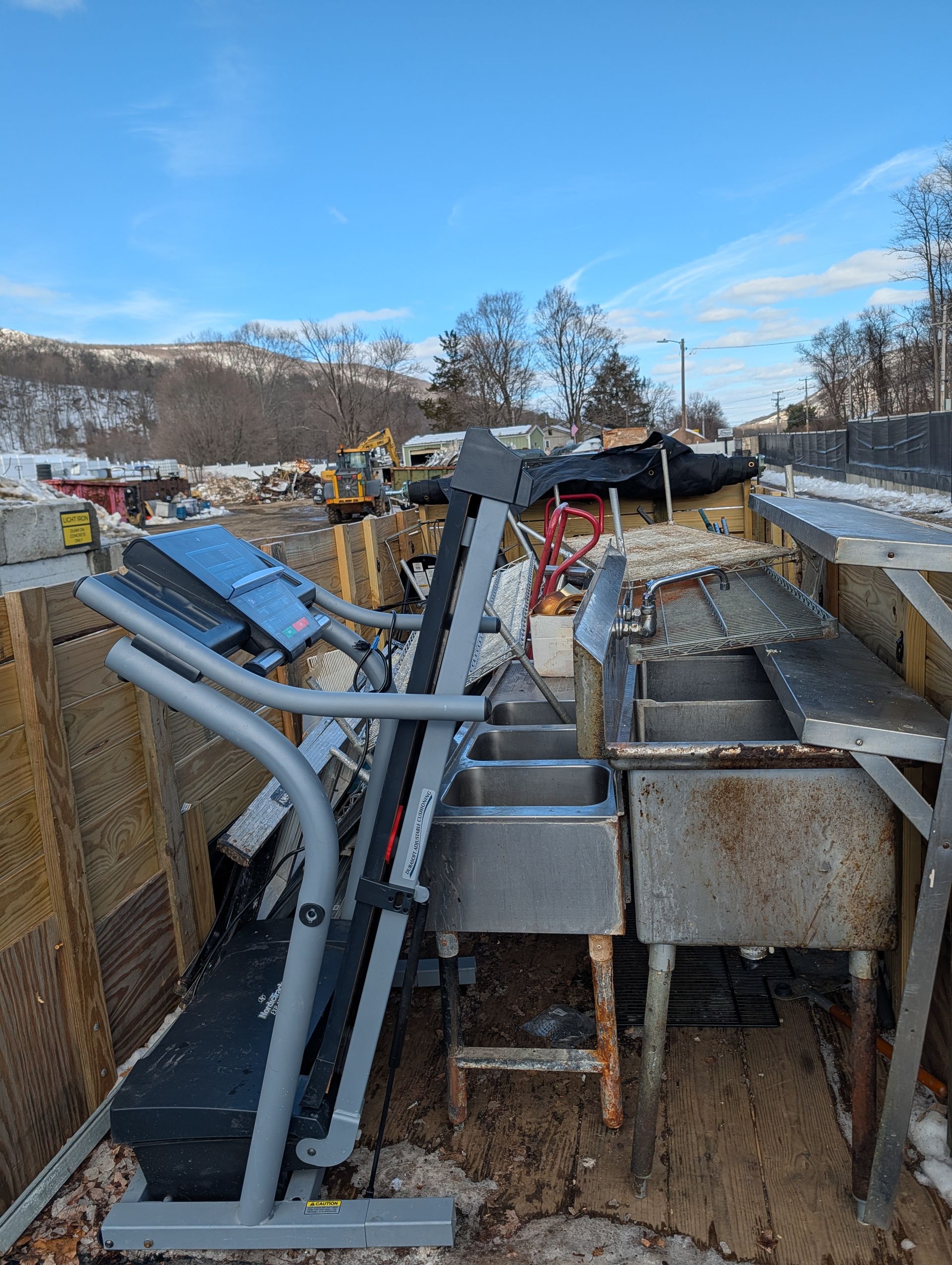 A folded treadmill and a triple sink in a debris-filled outdoor setting under a blue sky. A folded treadmill and a triple sink in a debris-filled outdoor setting under a blue sky.