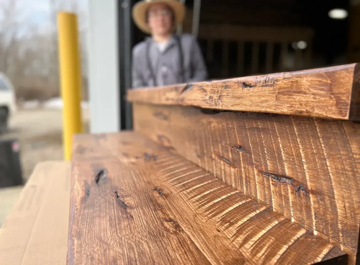 Stacked wooden beams in a workshop foreground, with a blurred person in a hat standing behind.