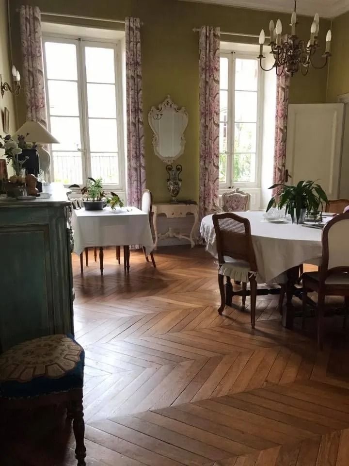 Elegant dining room with tall windows, herringbone wood floor, white tablecloths, and green-painted furniture