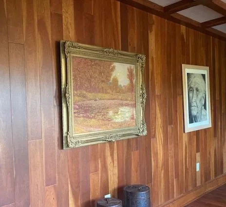 Wood-paneled wall with two framed artworks, one gold-framed landscape and one white-framed portrait.