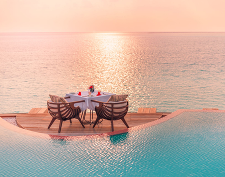 Table set for dinner on wooden deck over water, near infinity pool, at sunset.