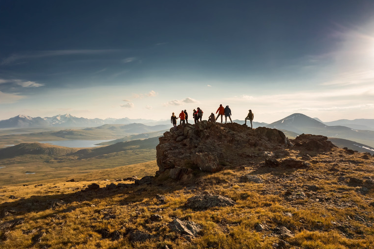 Hikers atop rocky peak with mountain views under a bright sky.
