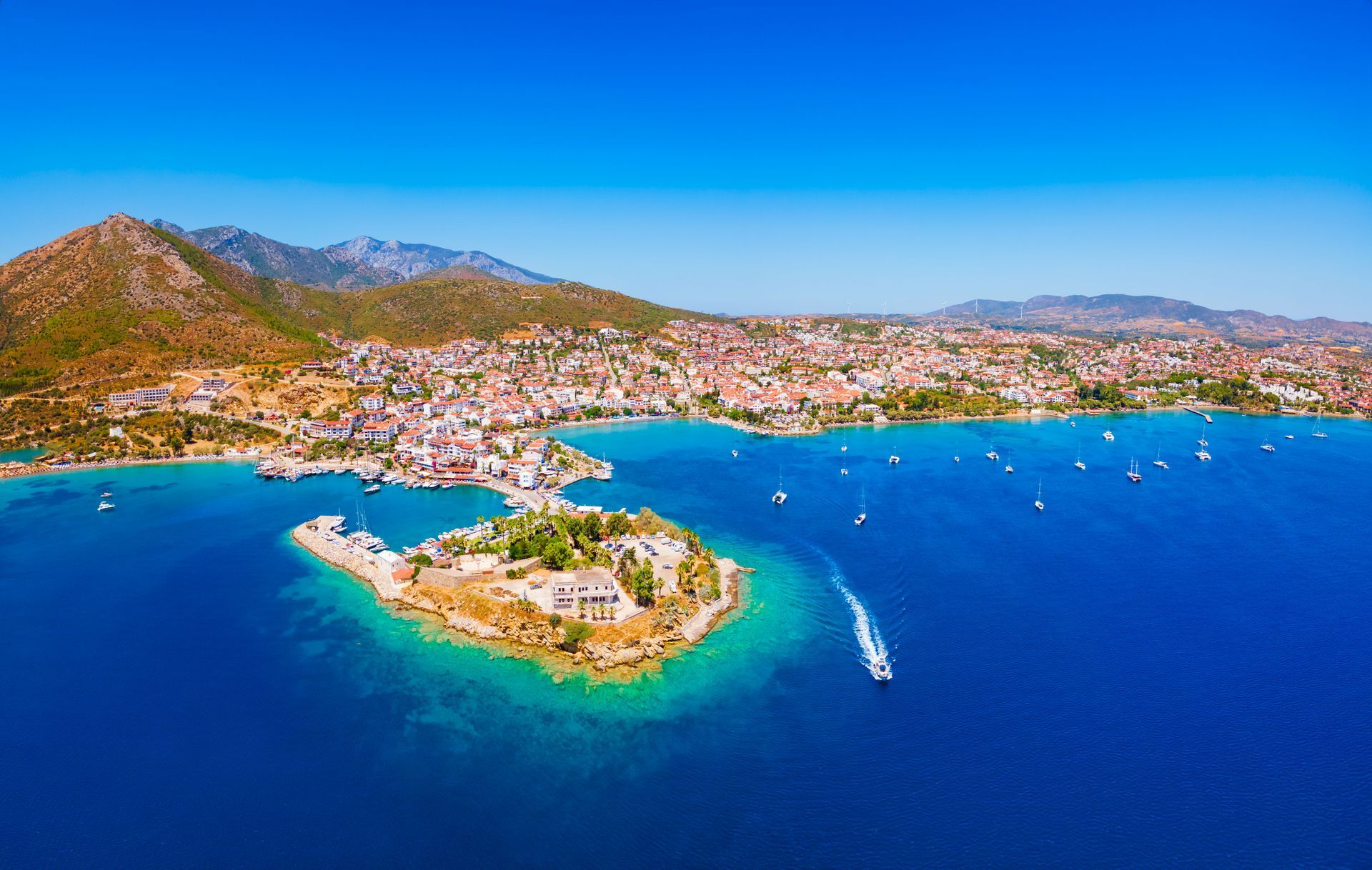 Aerial view of coastal town on bright blue sea, small island with ruins, mountains in the background.