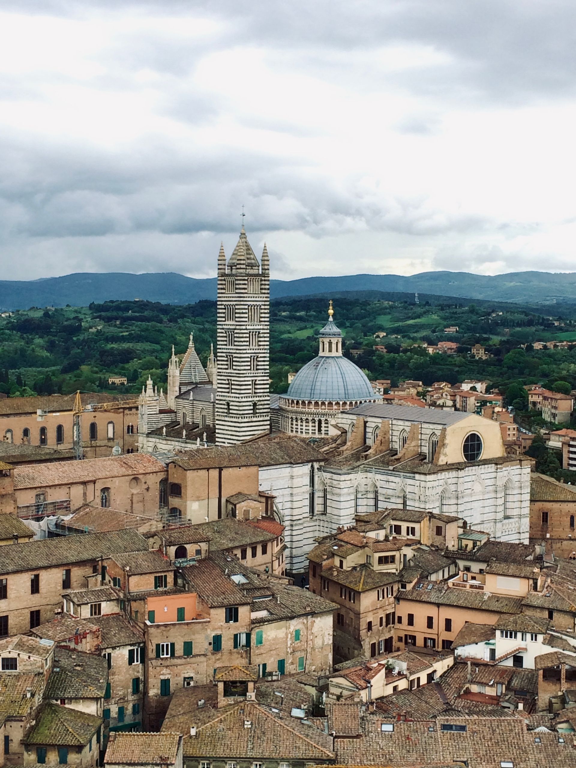 View of Siena, Italy, with striped bell tower, domed cathedral, and terracotta rooftops under cloudy sky.