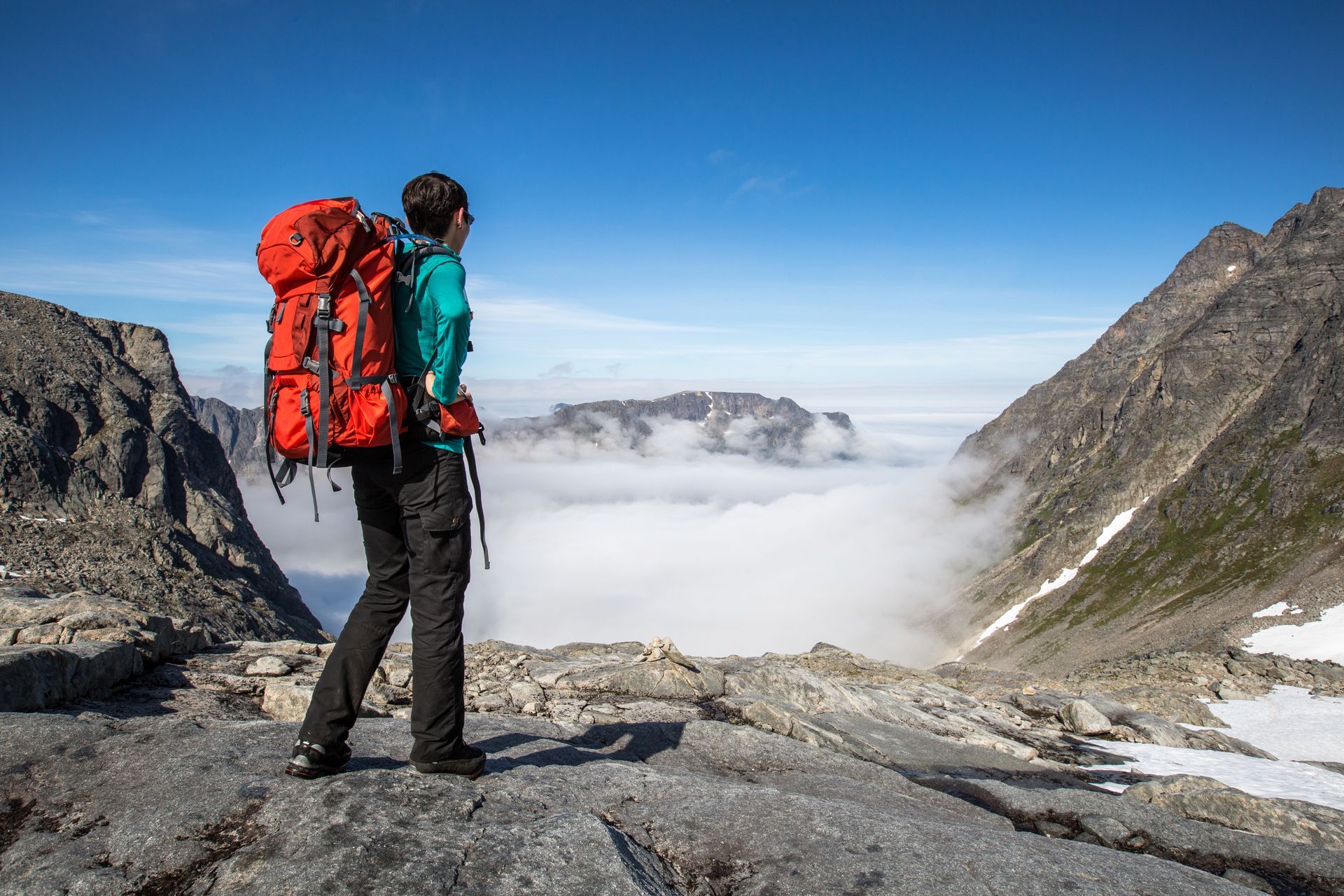 Person with red backpack looks at mountain vista, cloudy valley below.