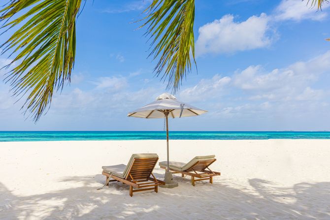White-sand tropical beach with two lounge chairs and umbrella under palm fronds, turquoise sea and blue sky