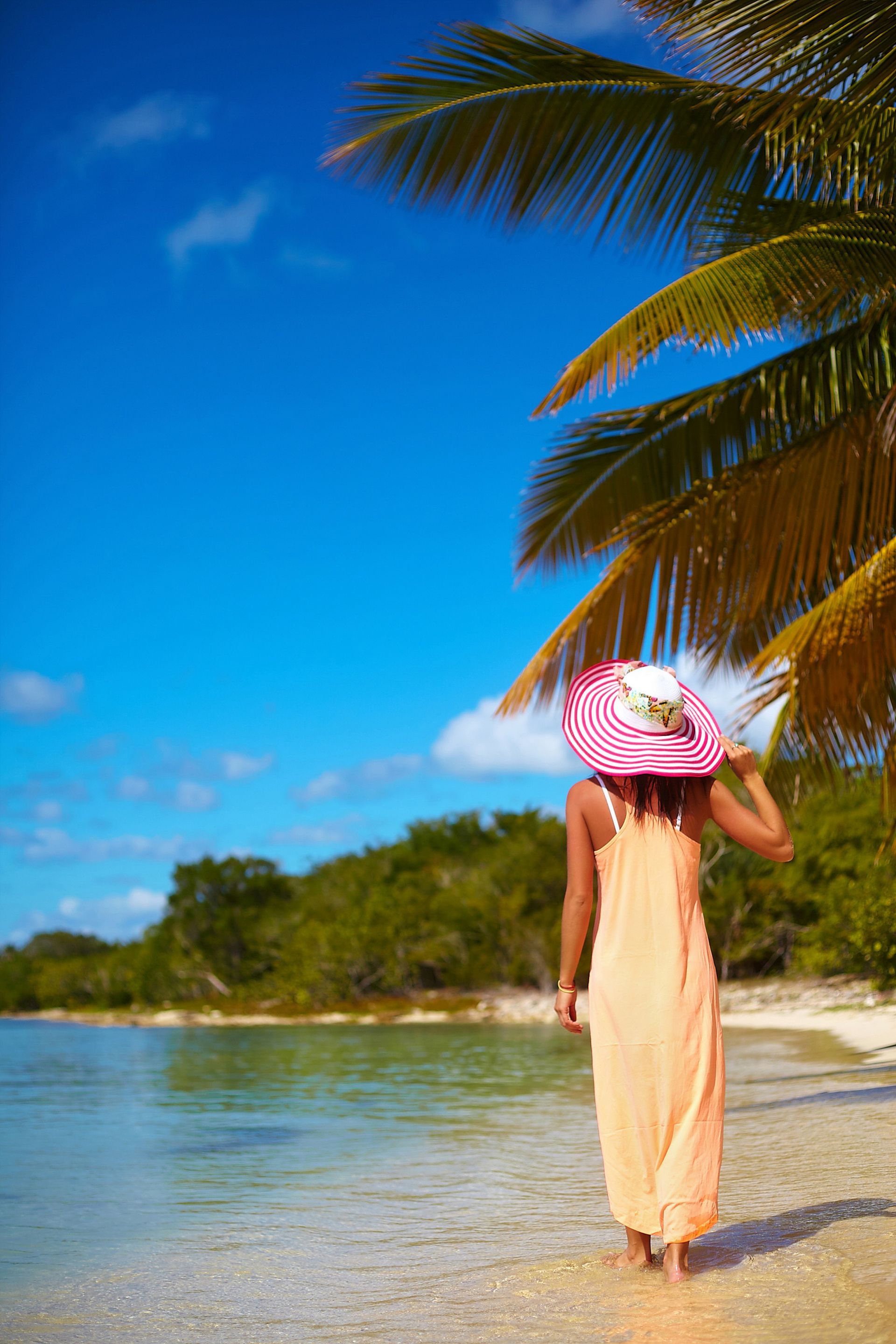 A person wearing a long peach dress and a large pink-and-white striped sun hat walks along a sandy tropical beach.