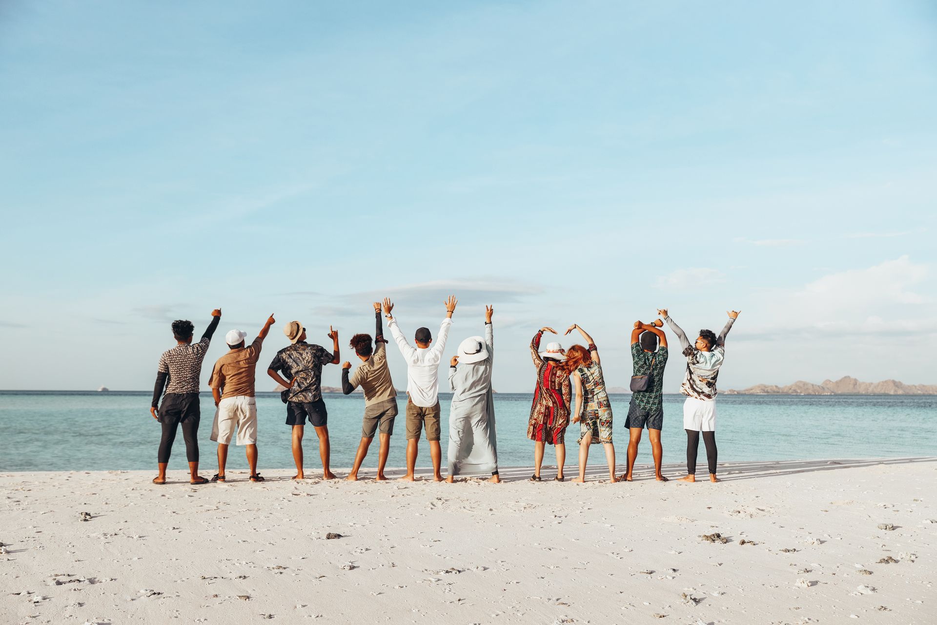 Group of people standing on a sandy beach, arms raised, facing the ocean under a clear blue sky.