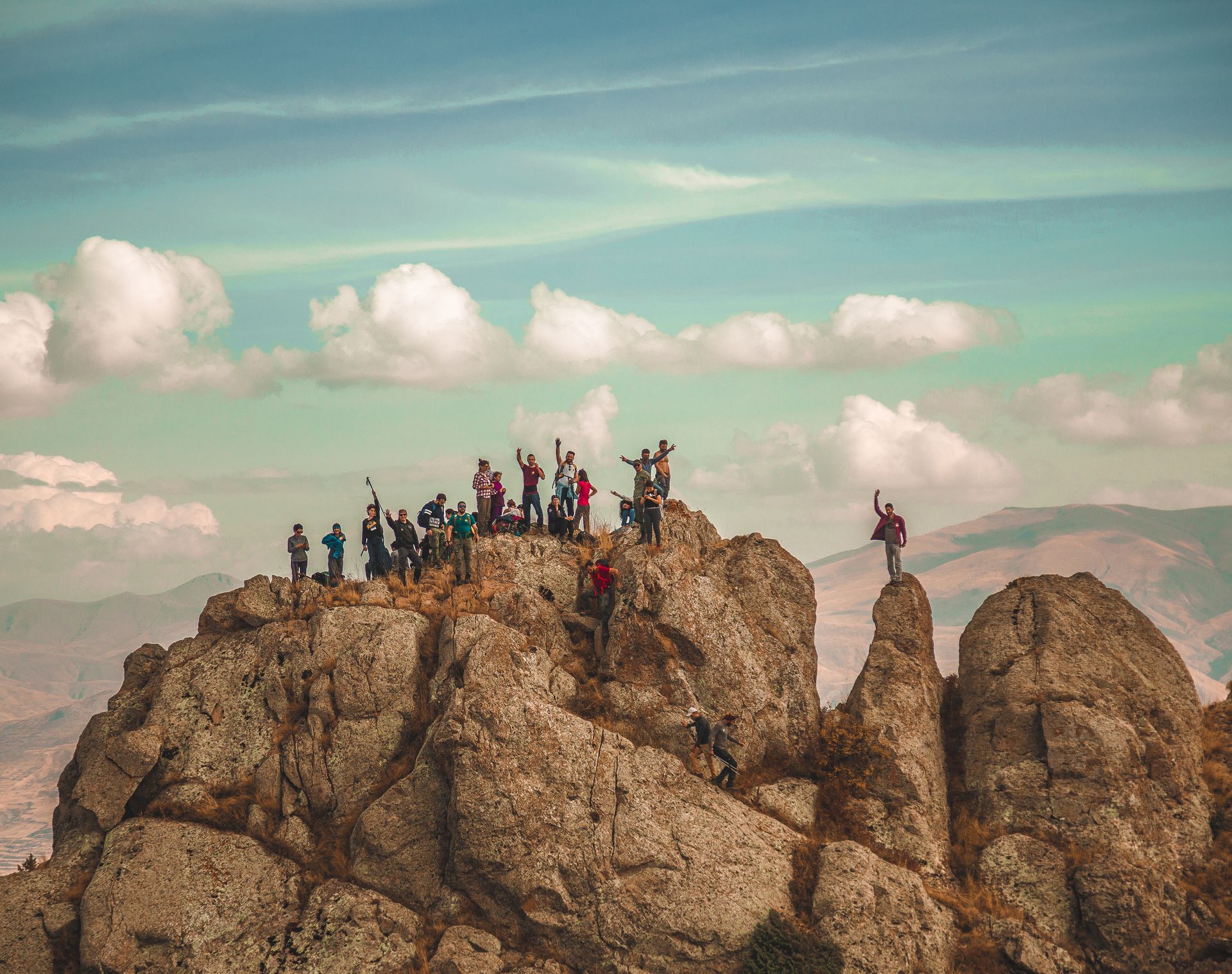 Group of people celebrating atop a rocky mountain peak under a cloudy blue sky.