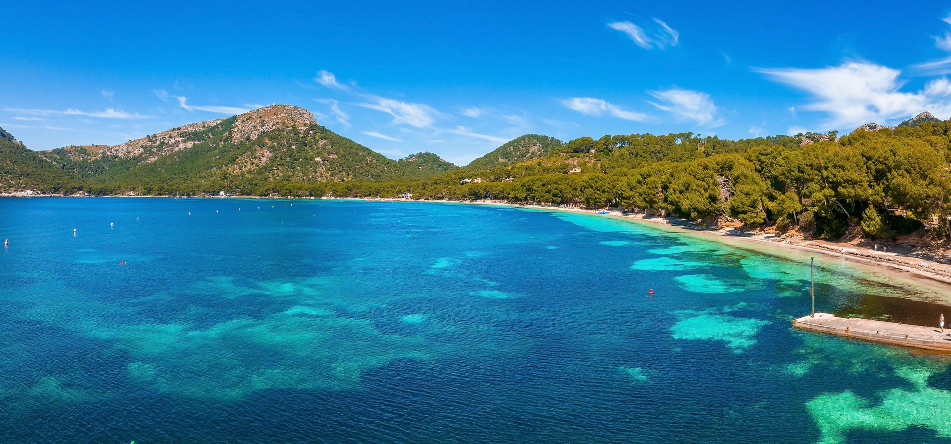 Bright blue ocean with clear turquoise shallows, green trees along the shore, and a mountainous backdrop under a blue sky.