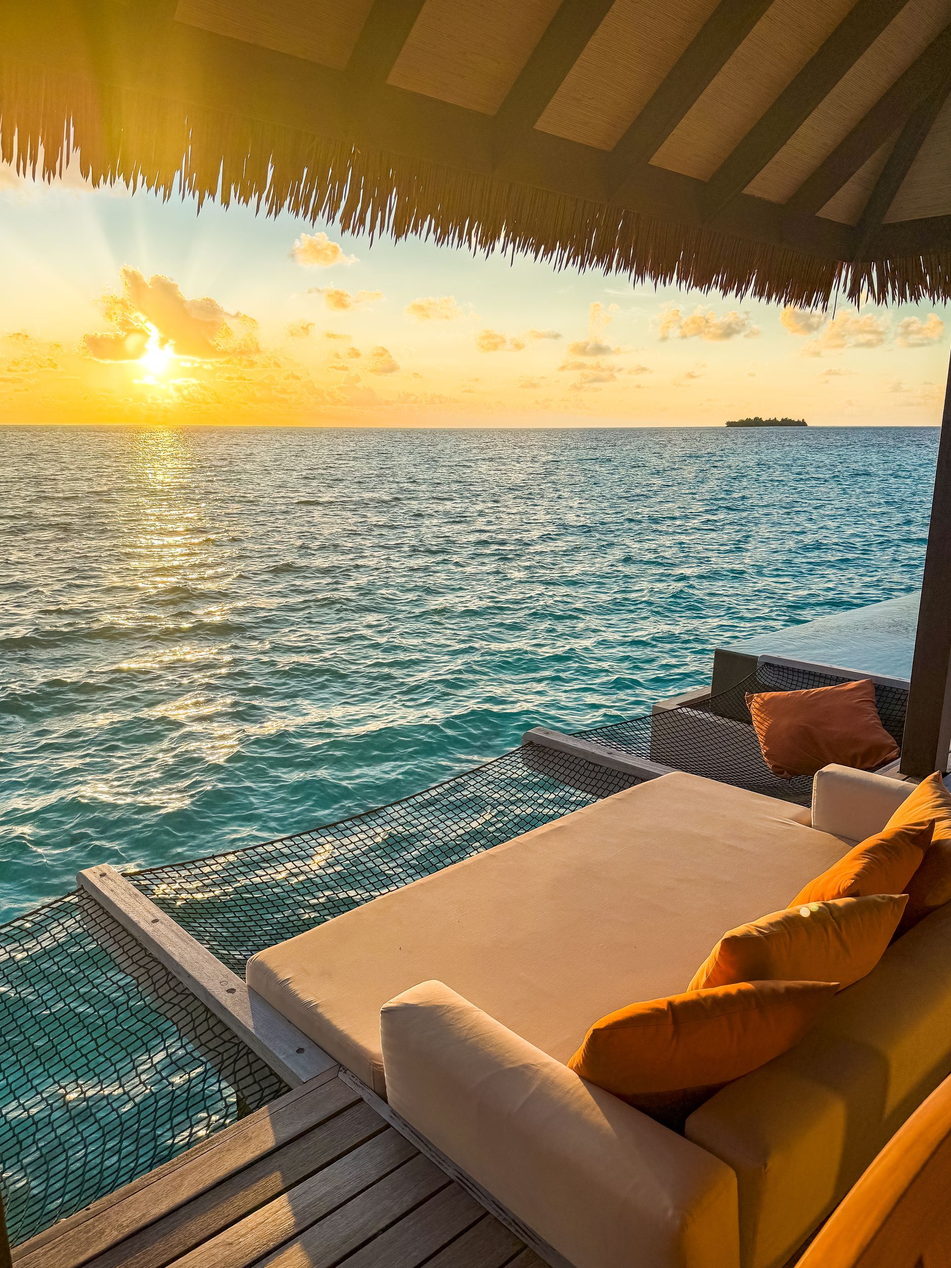 Ocean view with a lounge bed, sunset over the water, under a thatched roof.