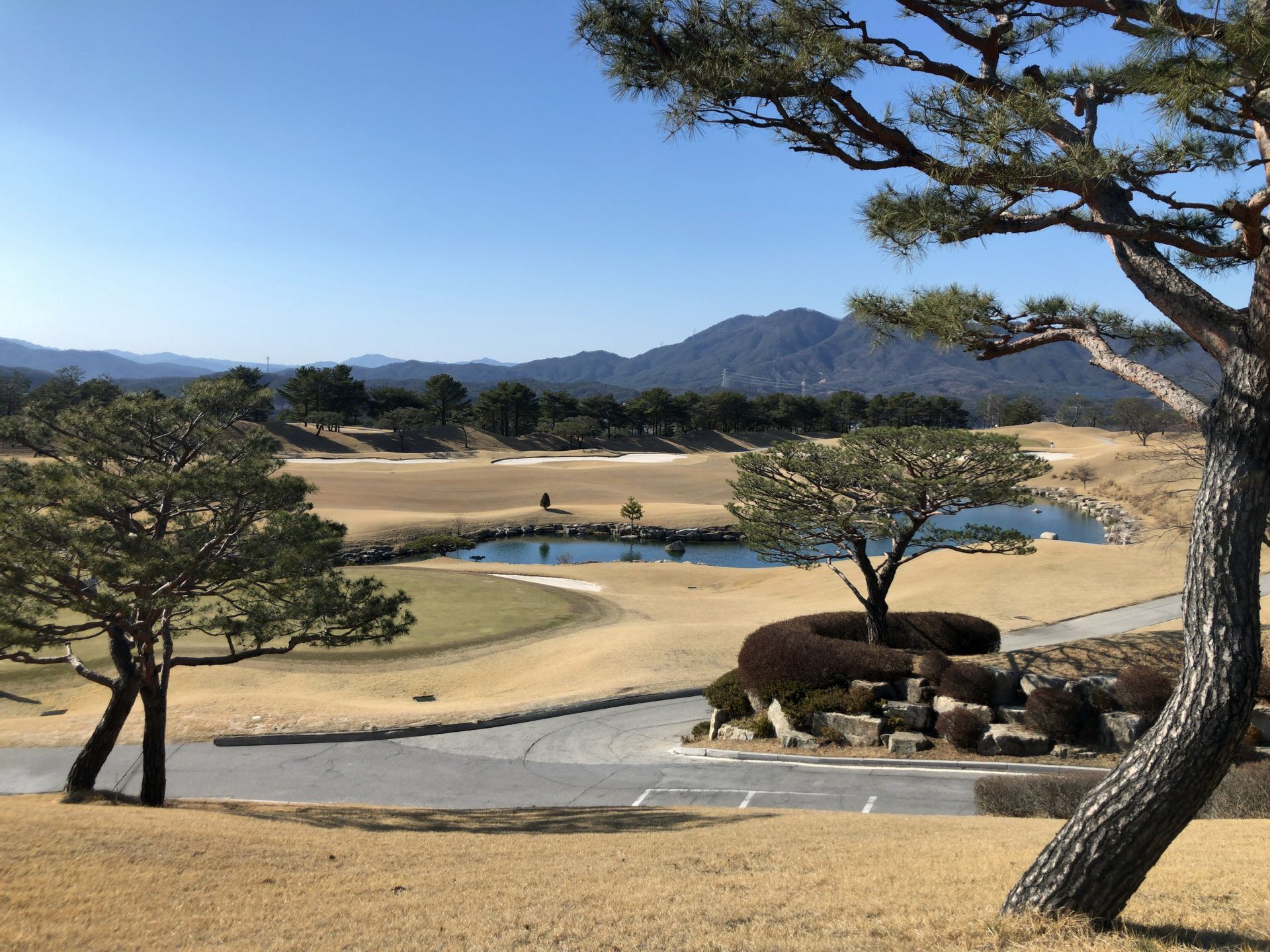 Golf course view with brown grass, trees, water feature, and distant mountains under a blue sky.