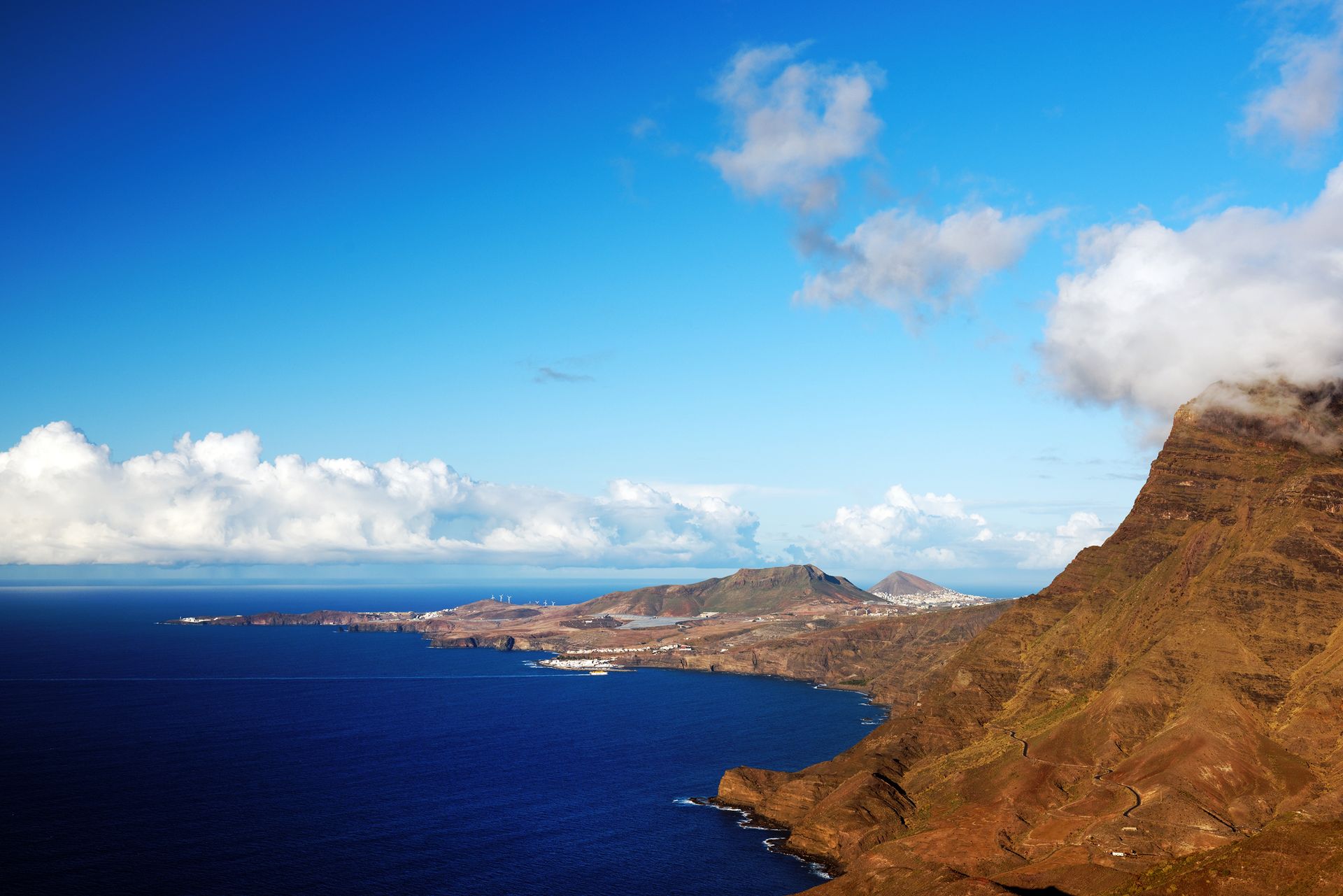 Blue ocean coastline under a bright blue sky with rocky cliffs and scattered white clouds.