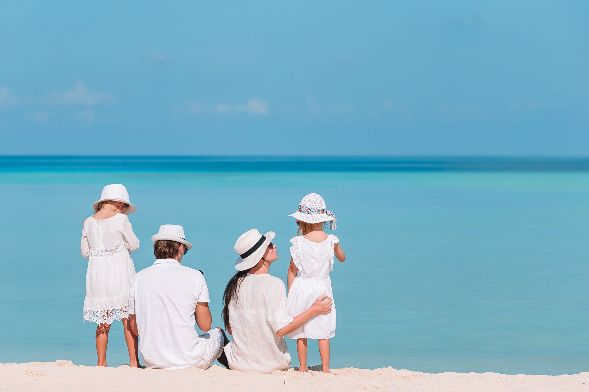 Family in white outfits sitting on a beach, looking at the ocean.