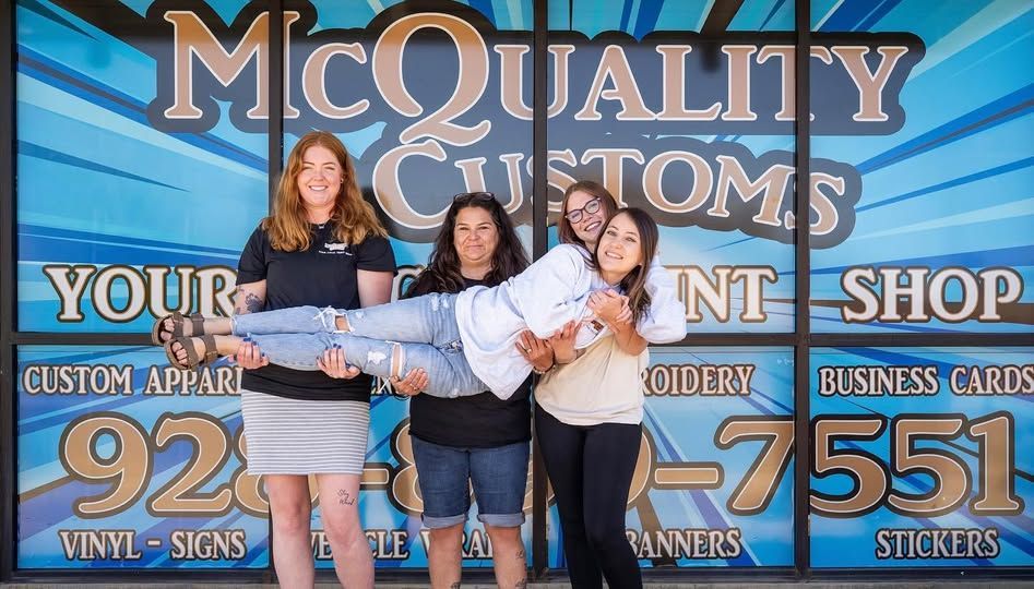 Four people posing in front of a shop. Two women are lifting a woman. The shop sign reads