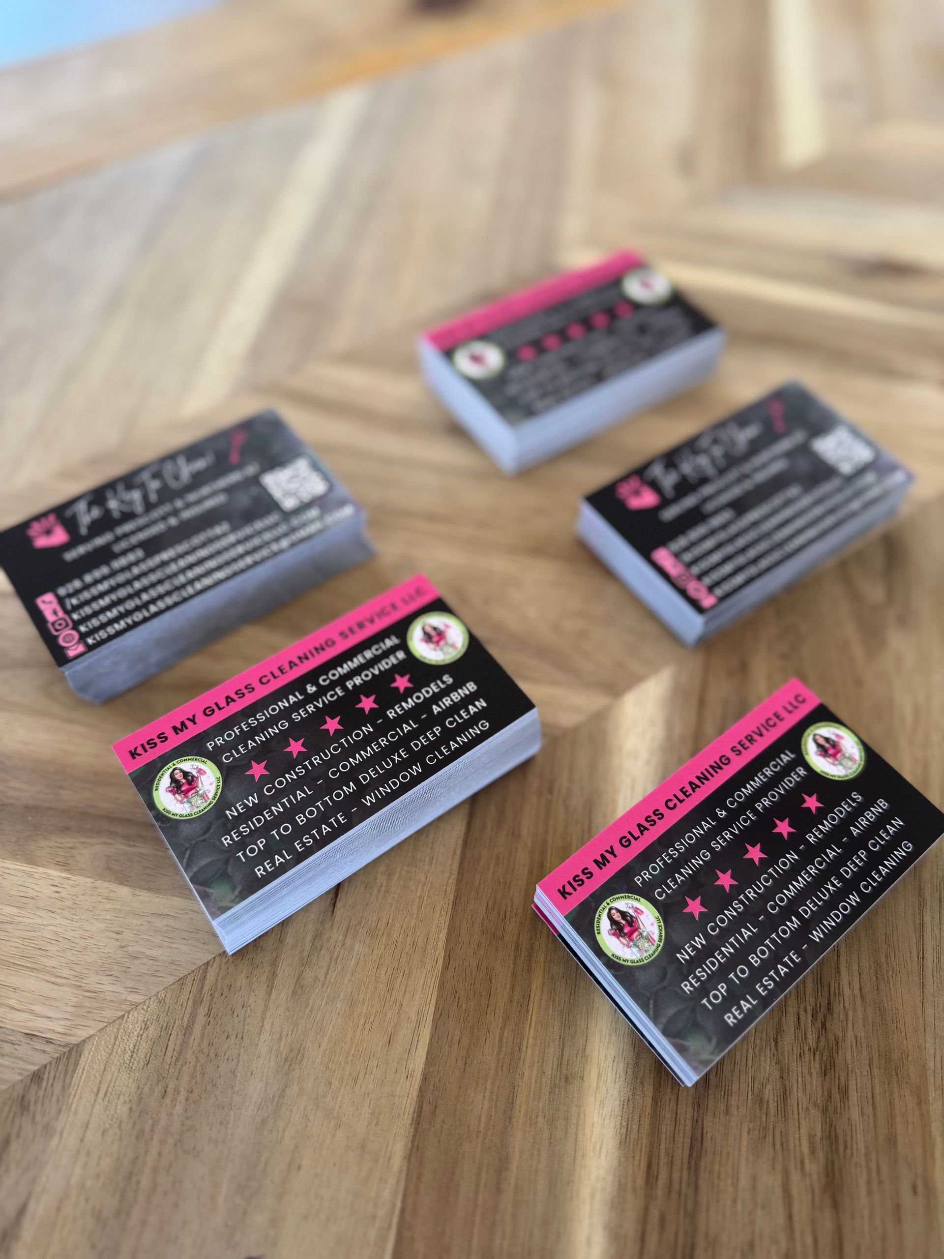 Five business cards with black backgrounds and pink accents are stacked on a wooden surface.