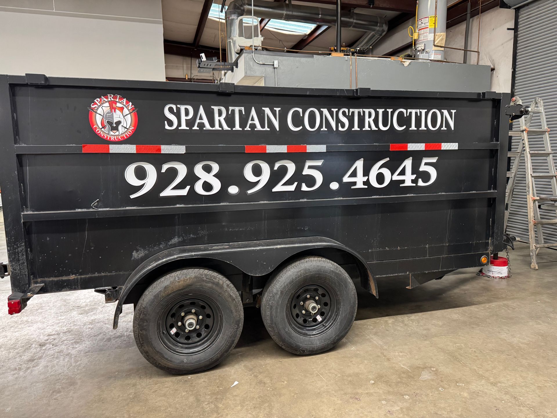 Black trailer for Spartan Construction with phone number 928.925.4645, black wheels, red and white reflective stripe.