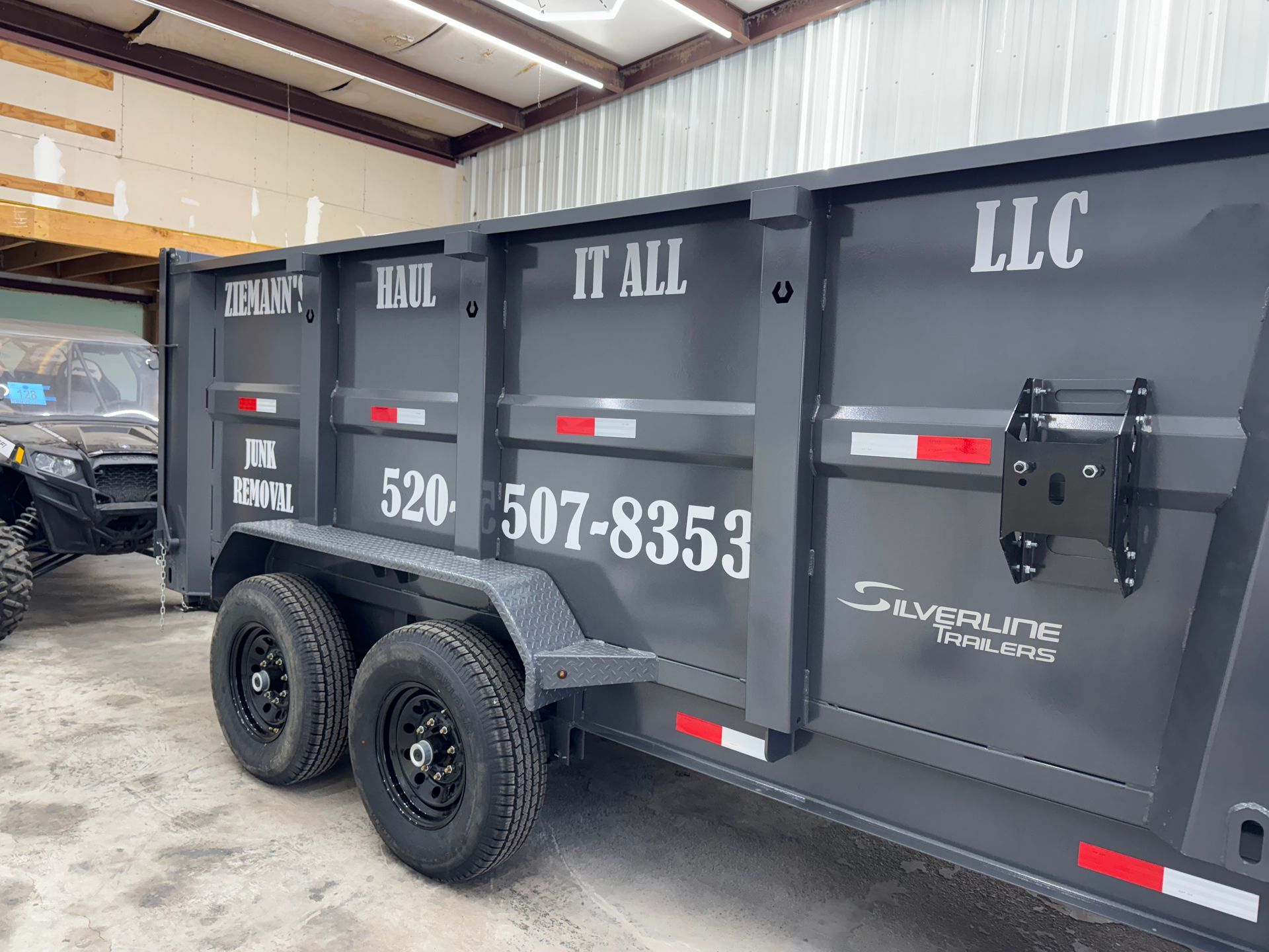 Dark gray dump trailer with black wheels, company logo, and phone number.