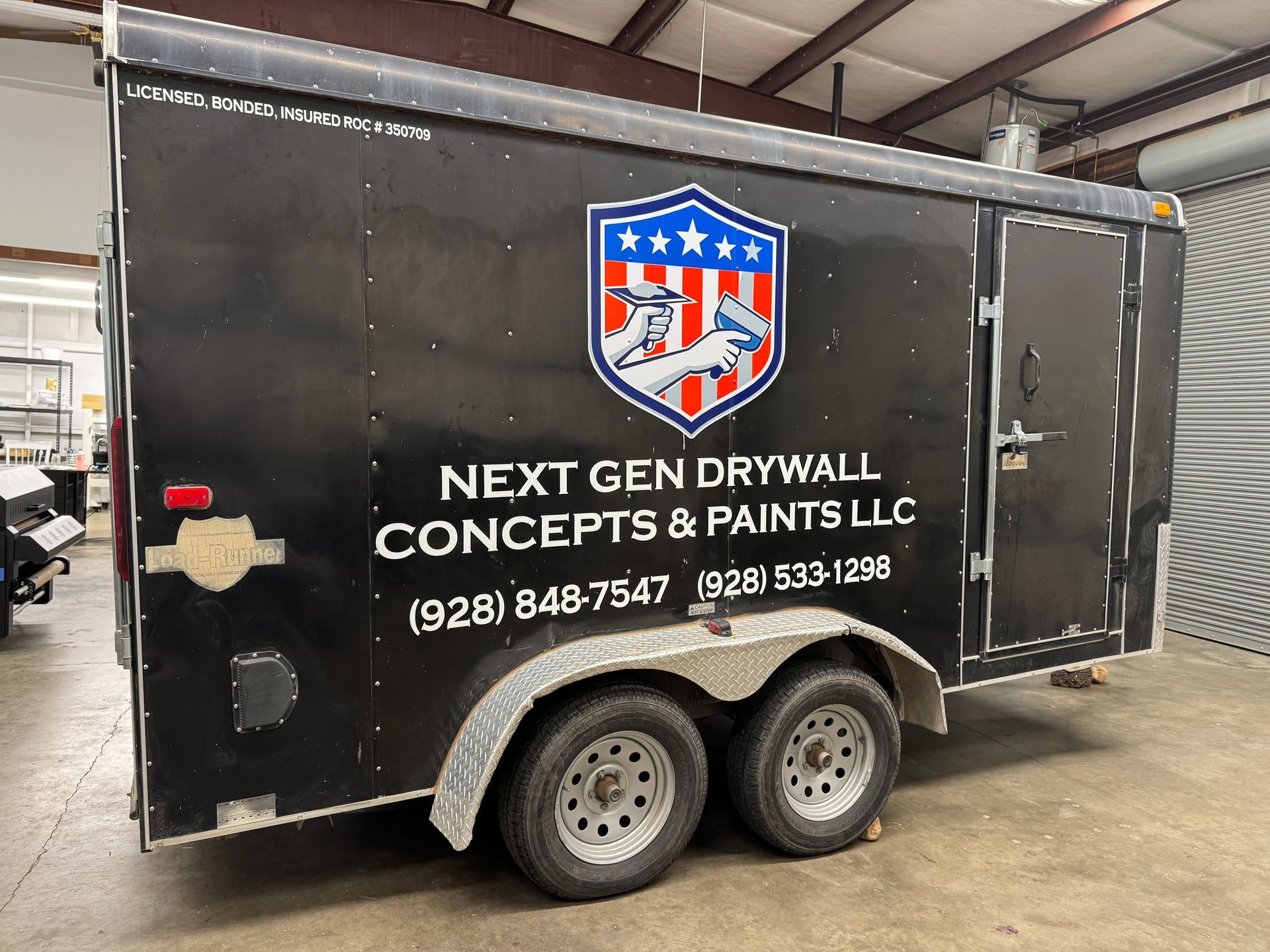 Black trailer with company logo and text: Next Gen Drywall Concepts & Paints LLC, including phone number, in a warehouse.