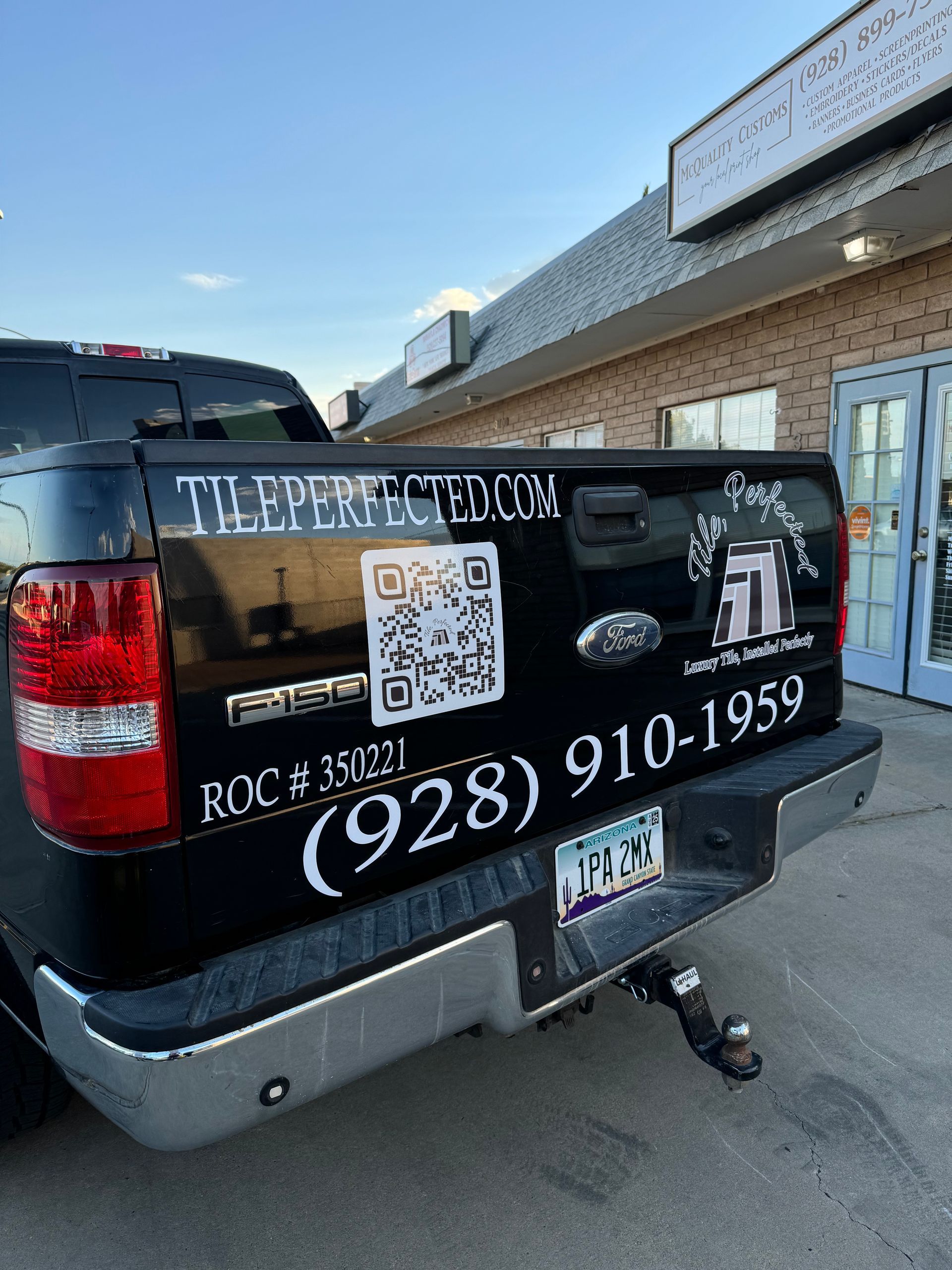 Black pickup truck advertising tileperfected.com, with a QR code and phone number in front of a brick building.