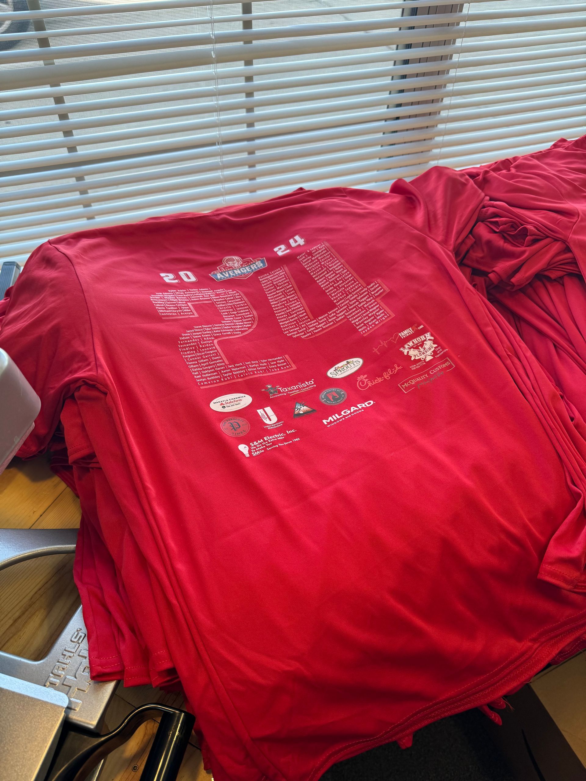 Red t-shirts stacked on a table. The top shirt has a design with white text and graphics. Sunlight streams through blinds.