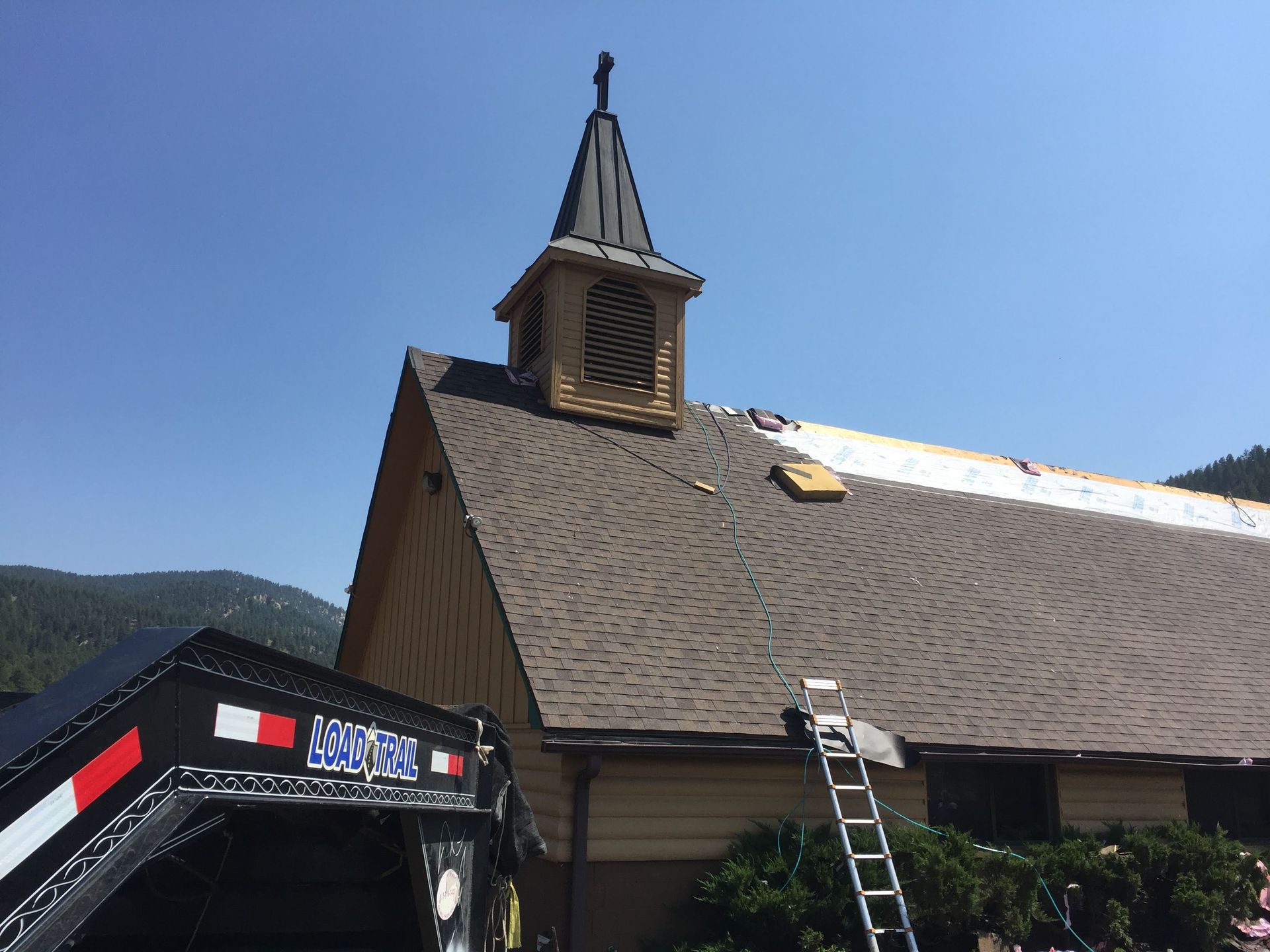 Church with workers replacing roof, brown shingles, steeple, trailer and ladder, mountains in background.