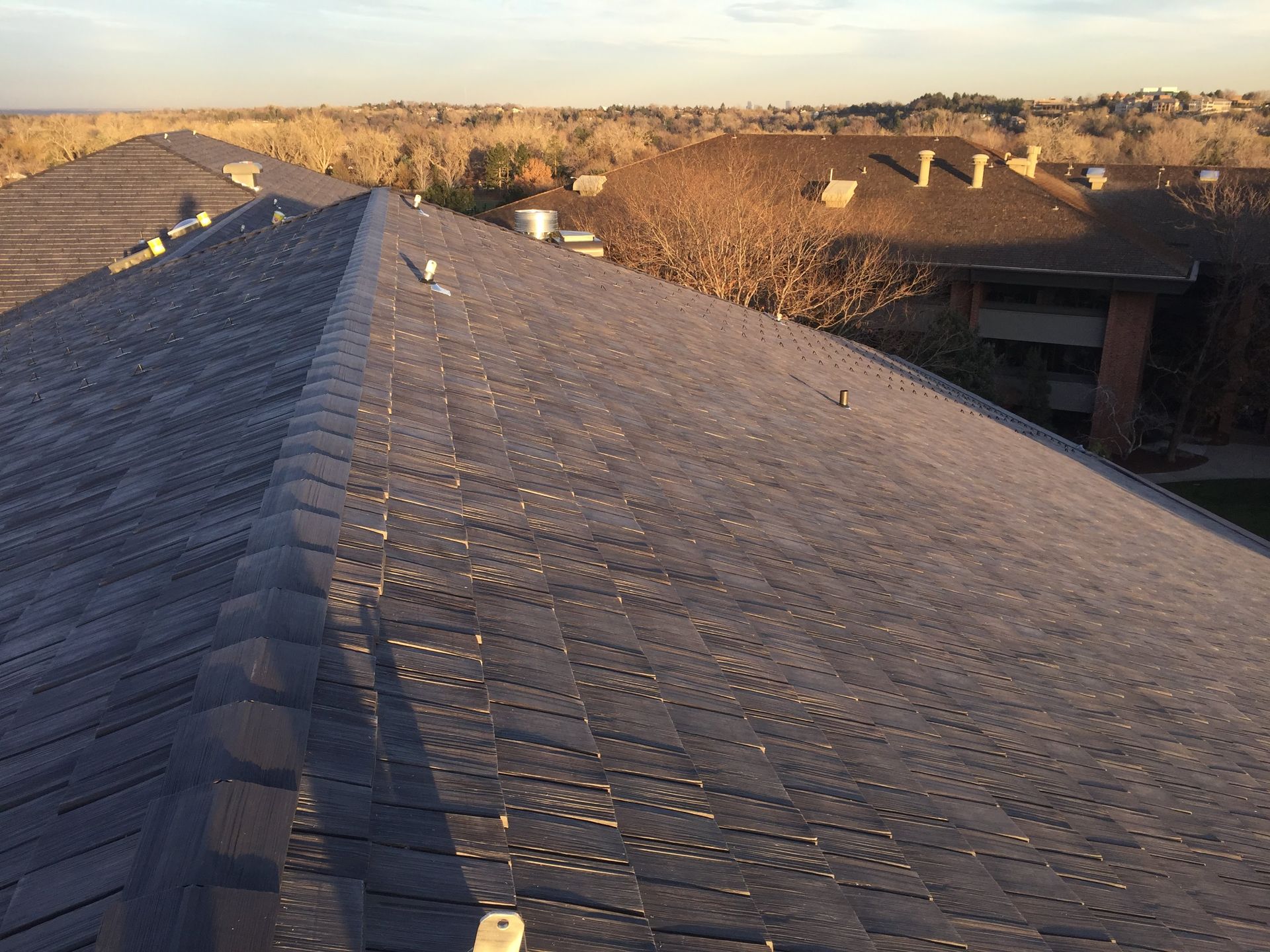 A gray asphalt shingle roof of a building with trees and houses in the background during daylight.