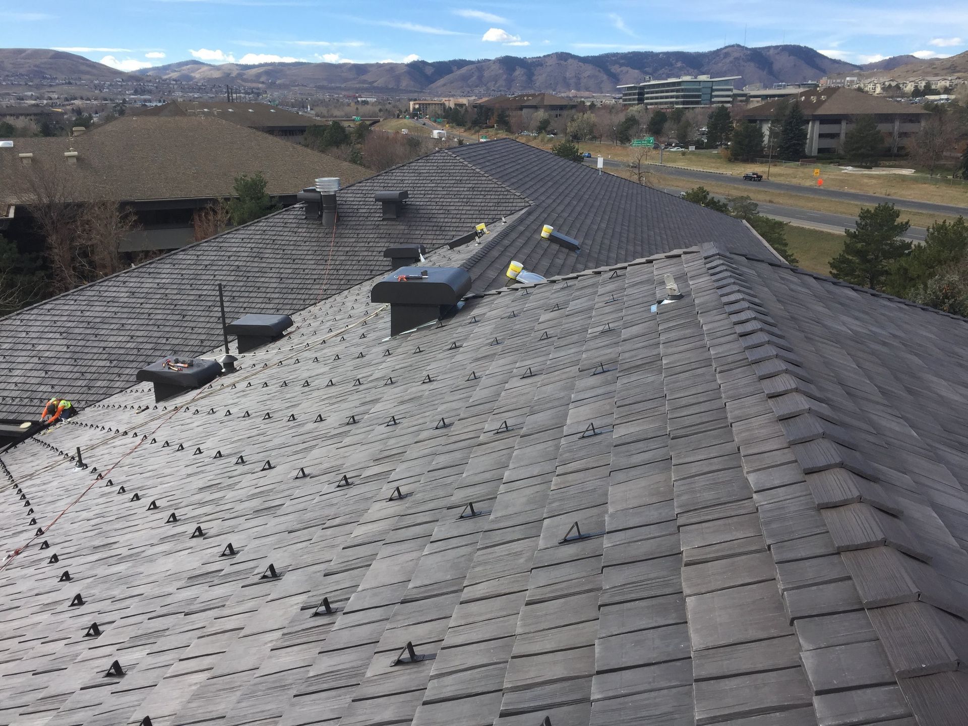 A rooftop covered in gray tiles with solar panel mounting hardware, set against a suburban landscape with distant mountains.