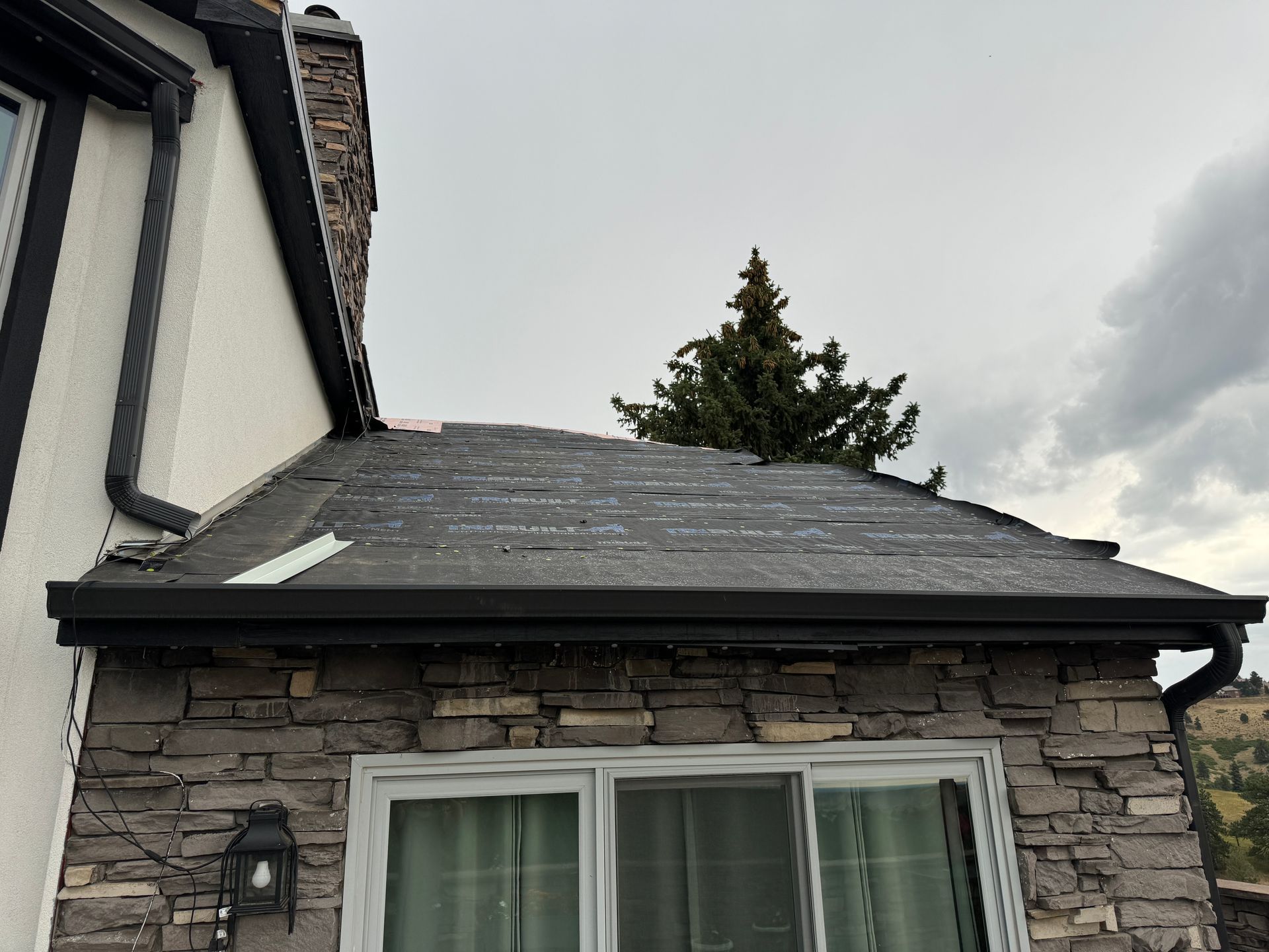 Close-up view of a roof with missing shingles, black gutters, and a stone wall beneath a cloudy sky.