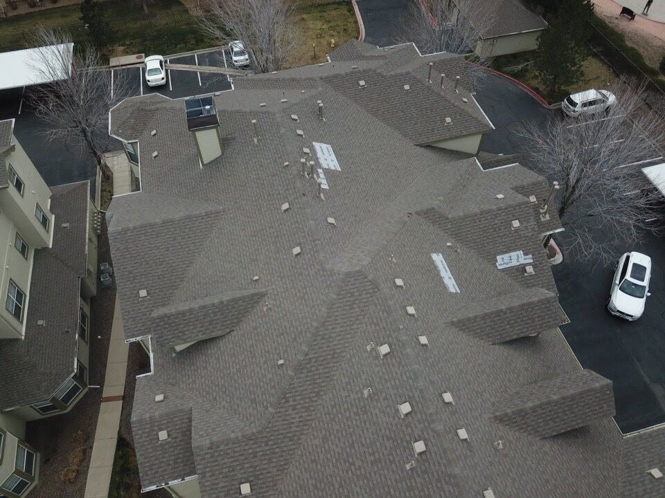 Aerial view of a multi-unit building with brown shingle roof, surrounded by parking and bare trees.