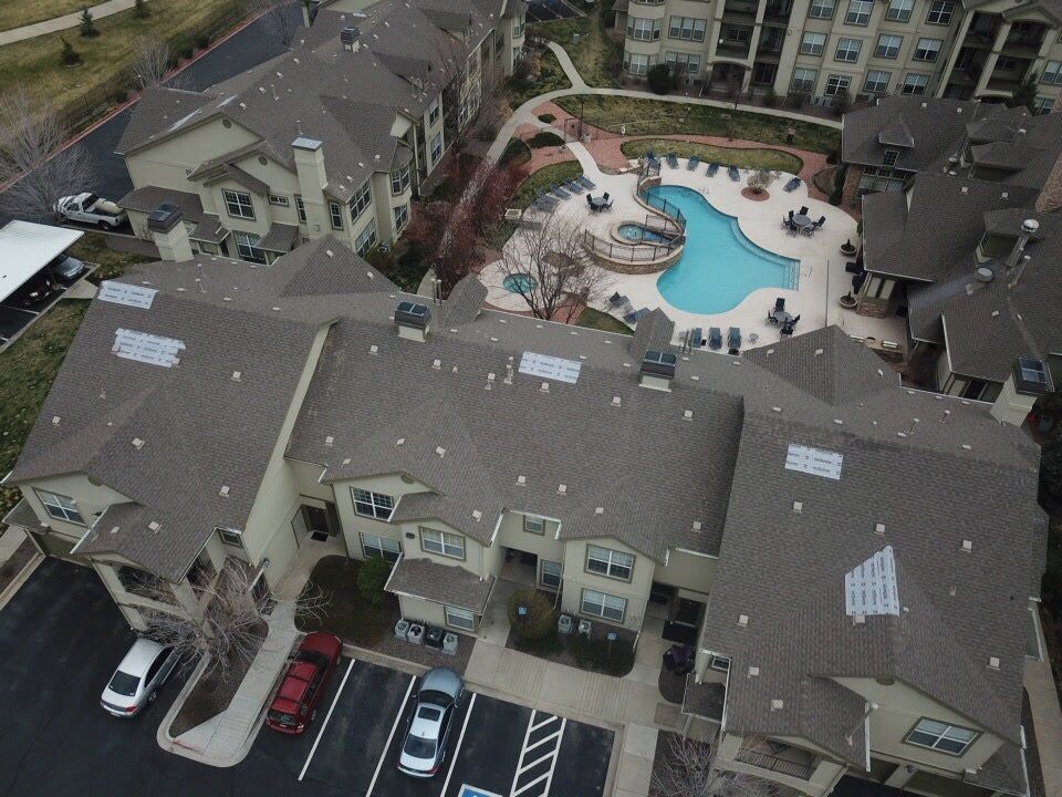 Aerial view of an apartment complex with a pool. Buildings have brown roofs. Cars parked in lot.