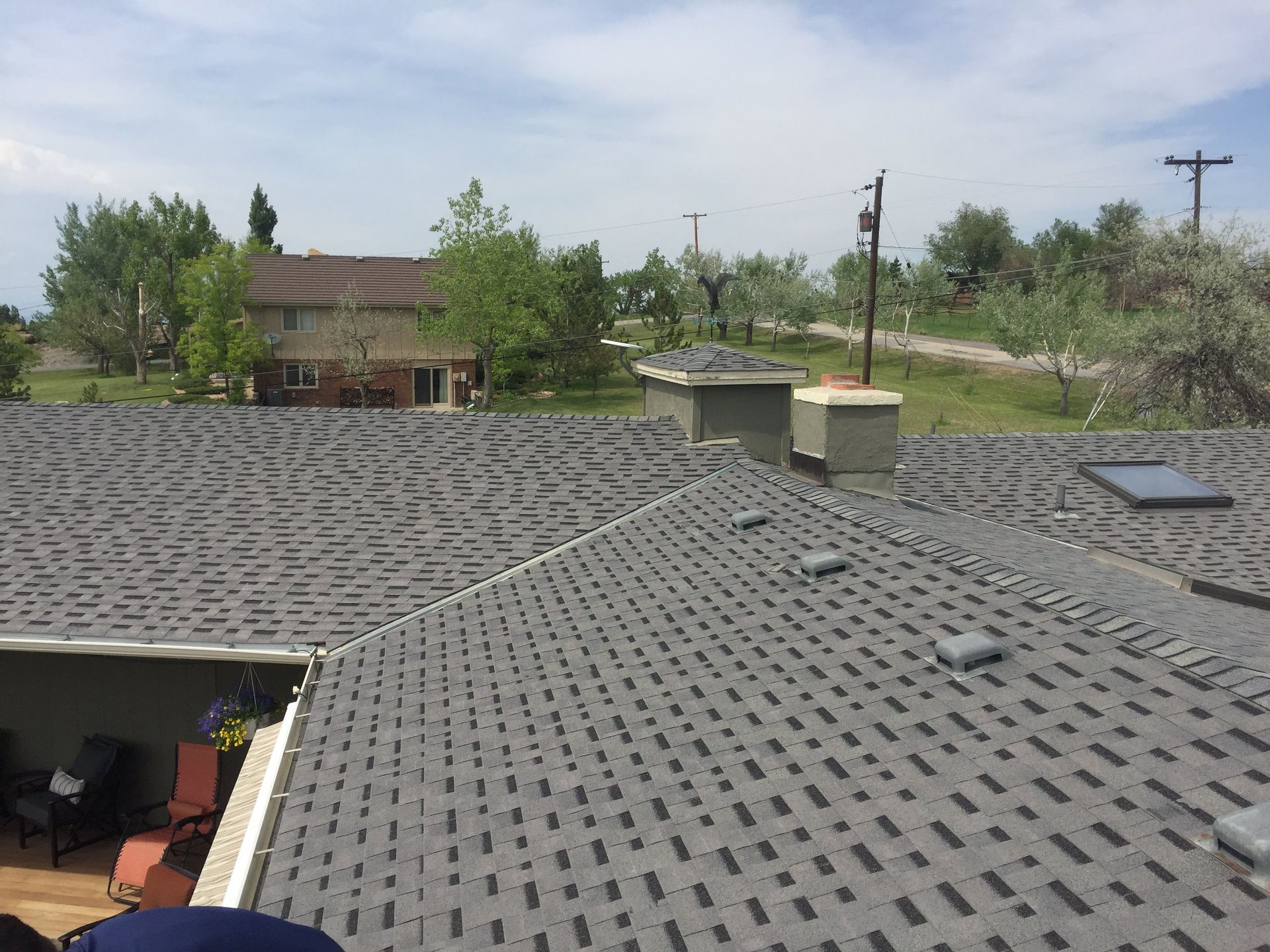 A dark gray shingle roof with a chimney and a distant house under a cloudy sky.