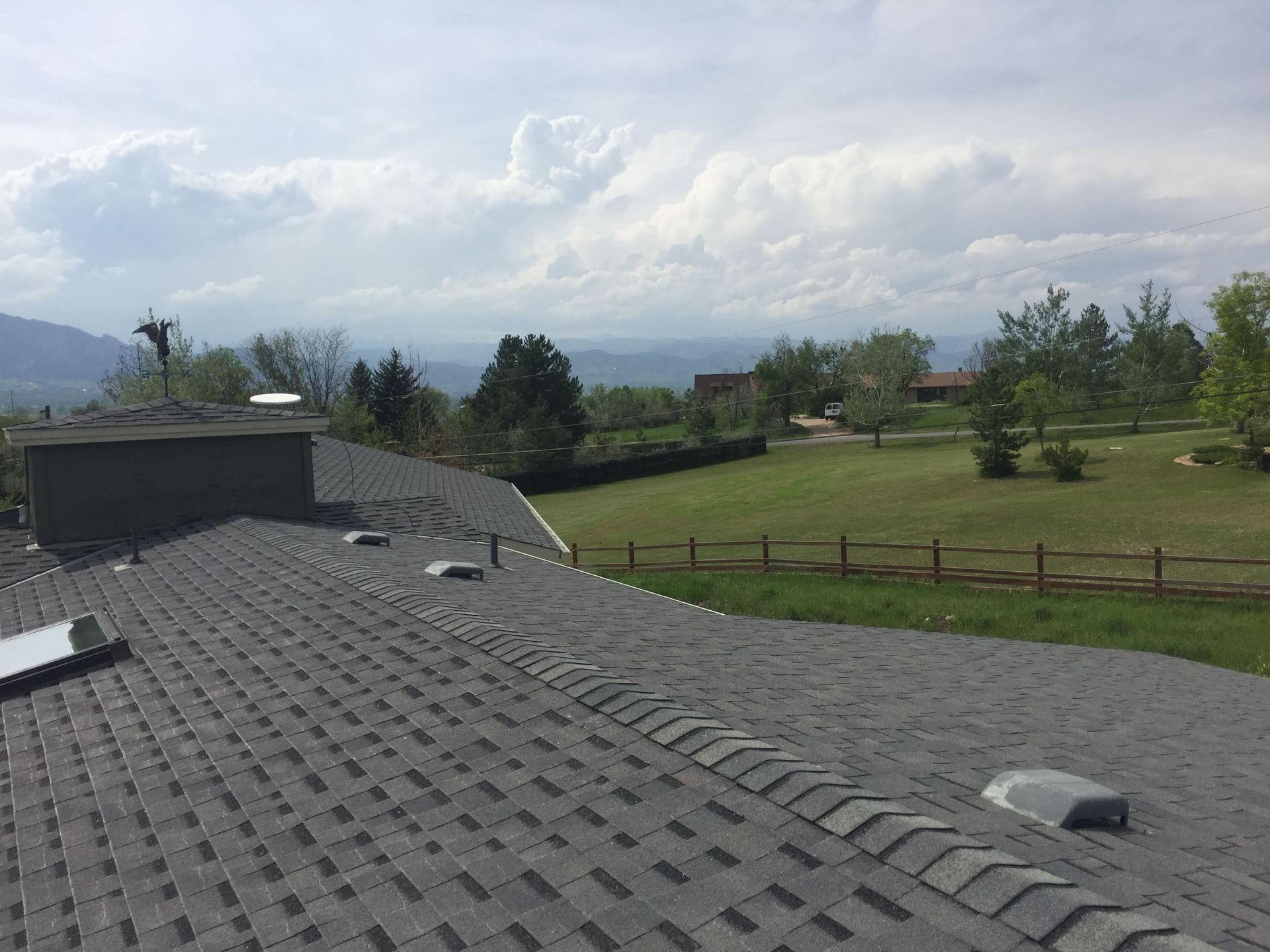 View from a dark shingle roof with vents over a green field, trees, and mountains under a cloudy sky.