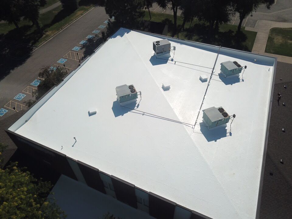 Aerial view of a white flat roof with HVAC units, brick building with parking and trees visible.